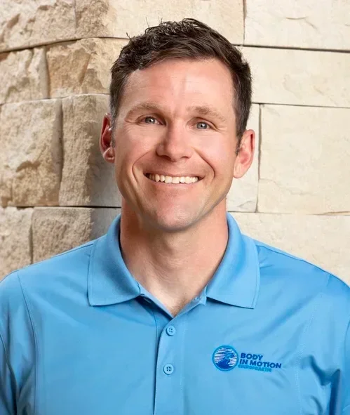 A smiling man with short dark hair wearing a blue collared shirt with a "Body In Motion" logo, standing outdoors against a stone wall.