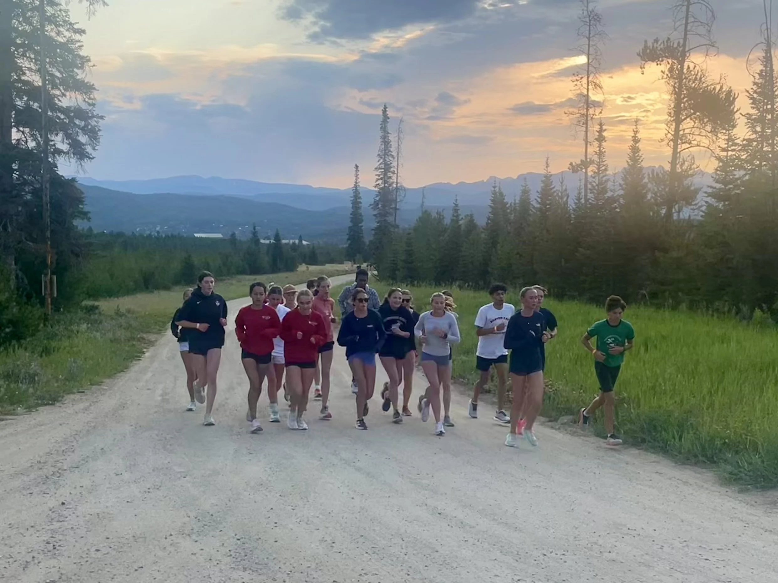 Group of people running on a dirt trail through a forested area at sunset with mountains in the background.