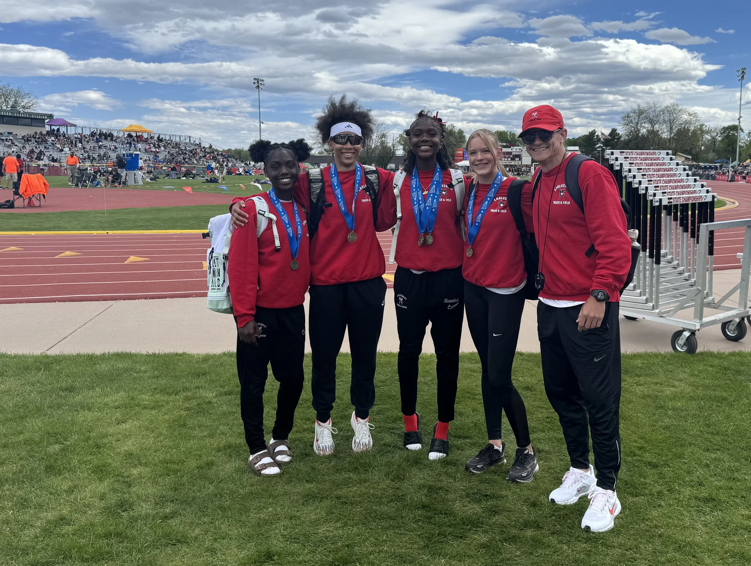 Five track athletes wearing red uniforms and medals, standing arm-in-arm on a grassy field at a stadium, with a track and audience in the background.