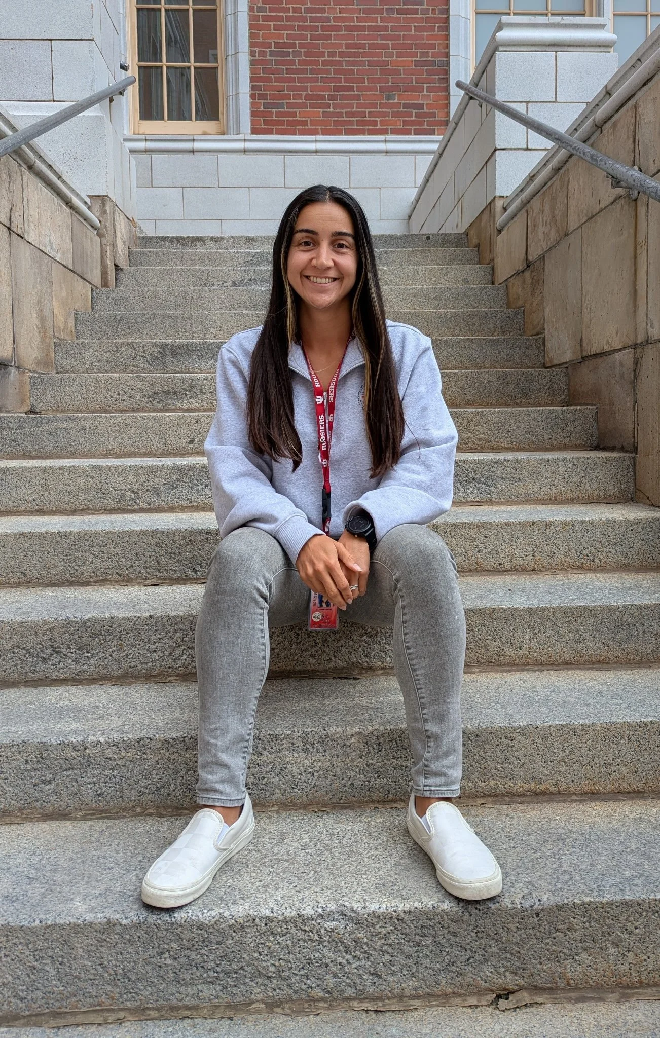 A young woman with long dark hair, sitting on outdoor concrete stairs, smiling, wearing a gray hoodie, gray jeans, white slip-on sneakers, and a red lanyard, with brick and stone building in the background.