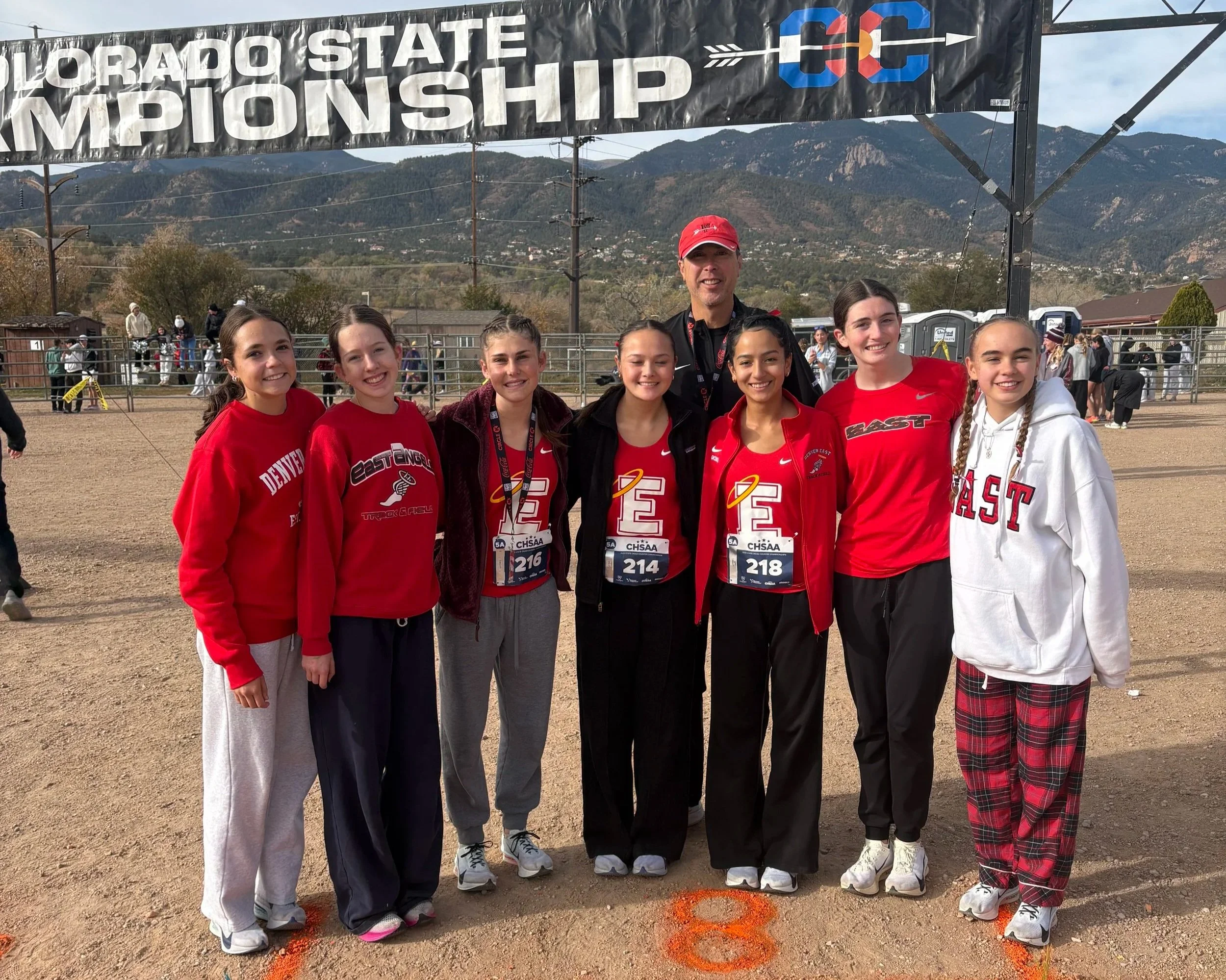 A group of seven young female track athletes and a male coach, standing on a dirt field after a race at the Colorado State Championship, with mountains in the background. They are smiling, wearing red and white track uniforms with race bibs, and are under a large banner that reads 'Colorado State Championship'.