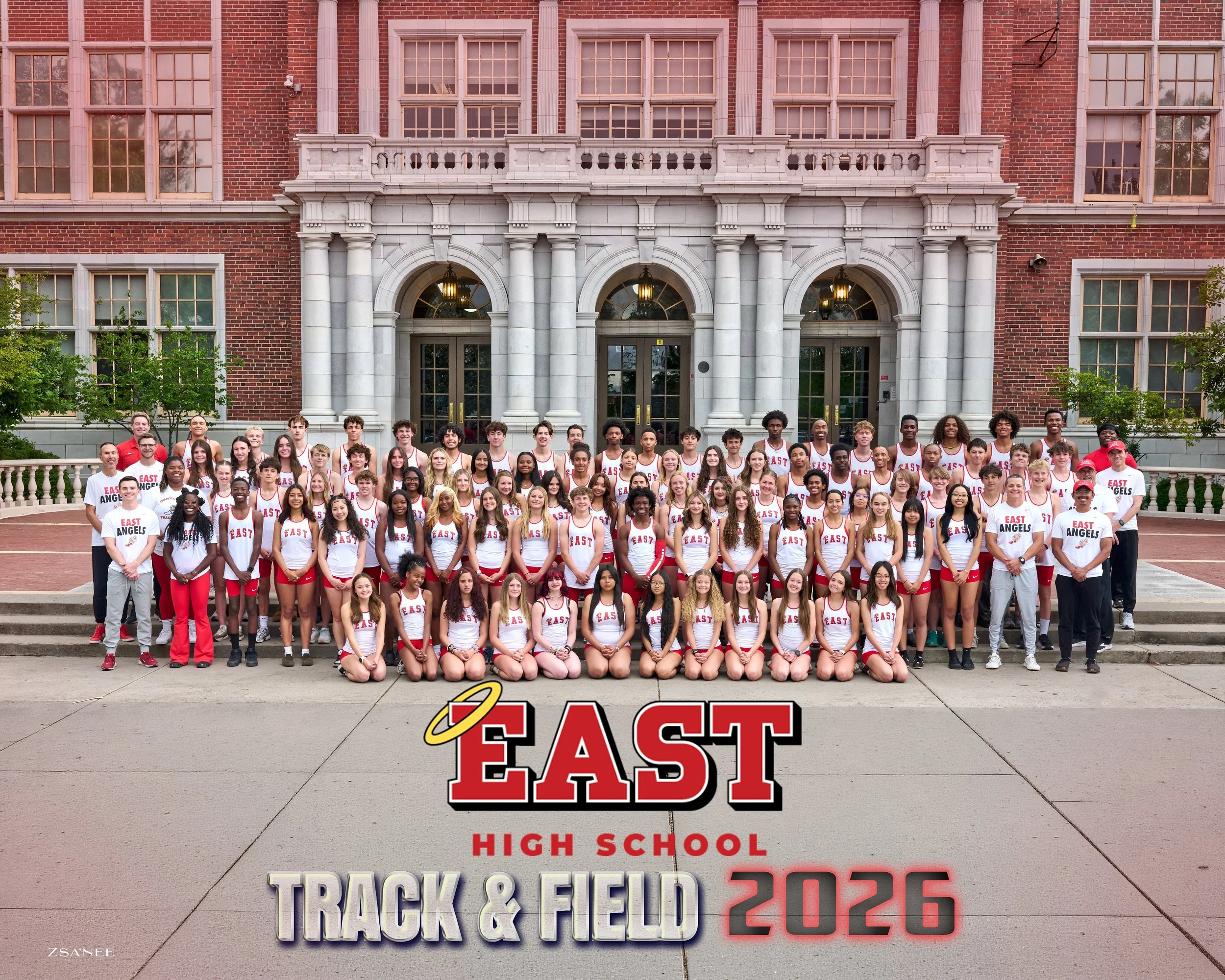 A group of students and coaches in front of a red brick high school building, wearing athletic uniforms that say East Angels. The group appears to be gathered for a track and field event, with a large sign on the sidewalk displaying 'EAST HIGH SCHOOL TRACK & FIELD 2023'.