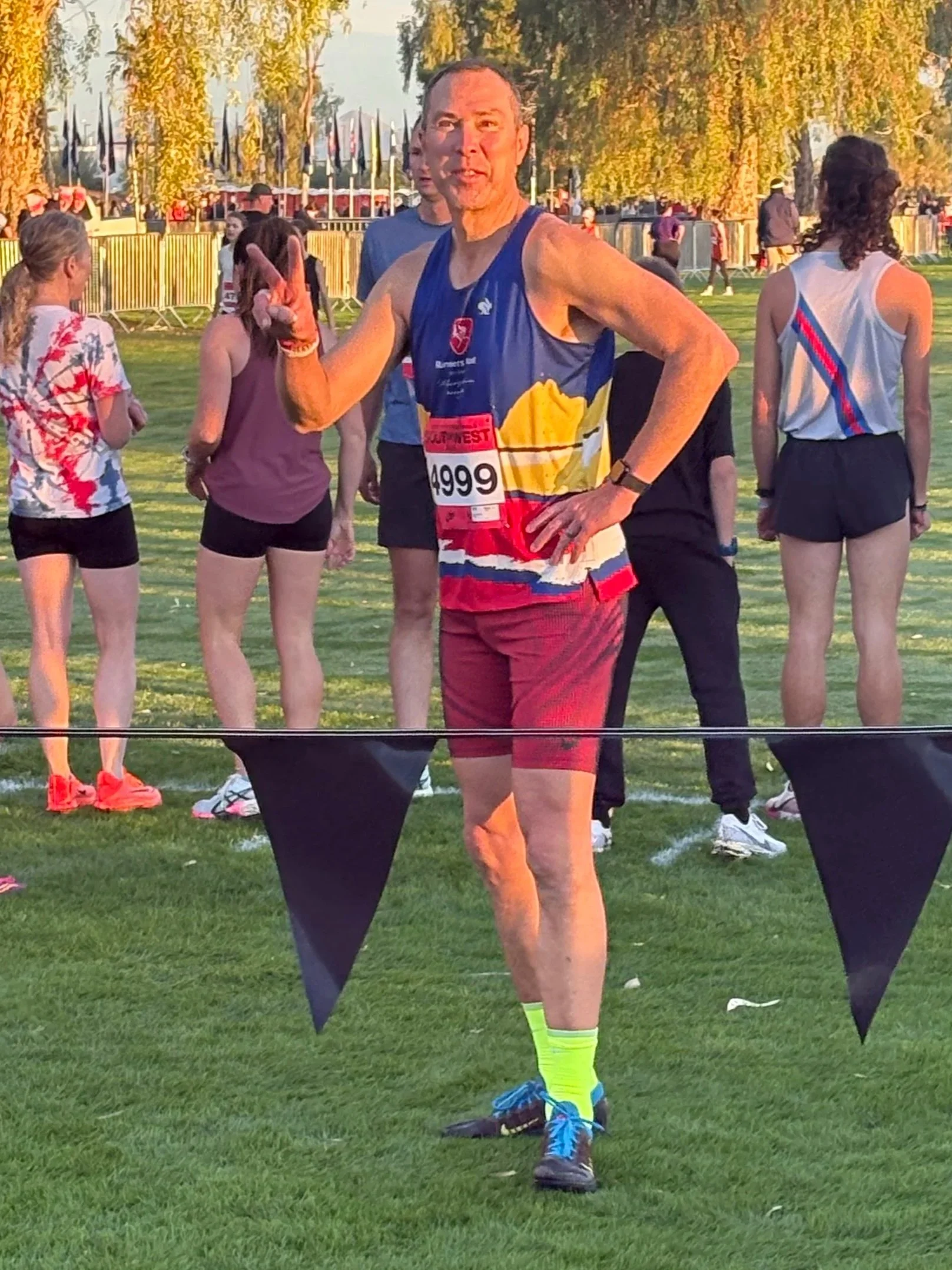 An older man in athletic wear making a peace sign during a running event at an outdoor track or park, with several other runners in the background.