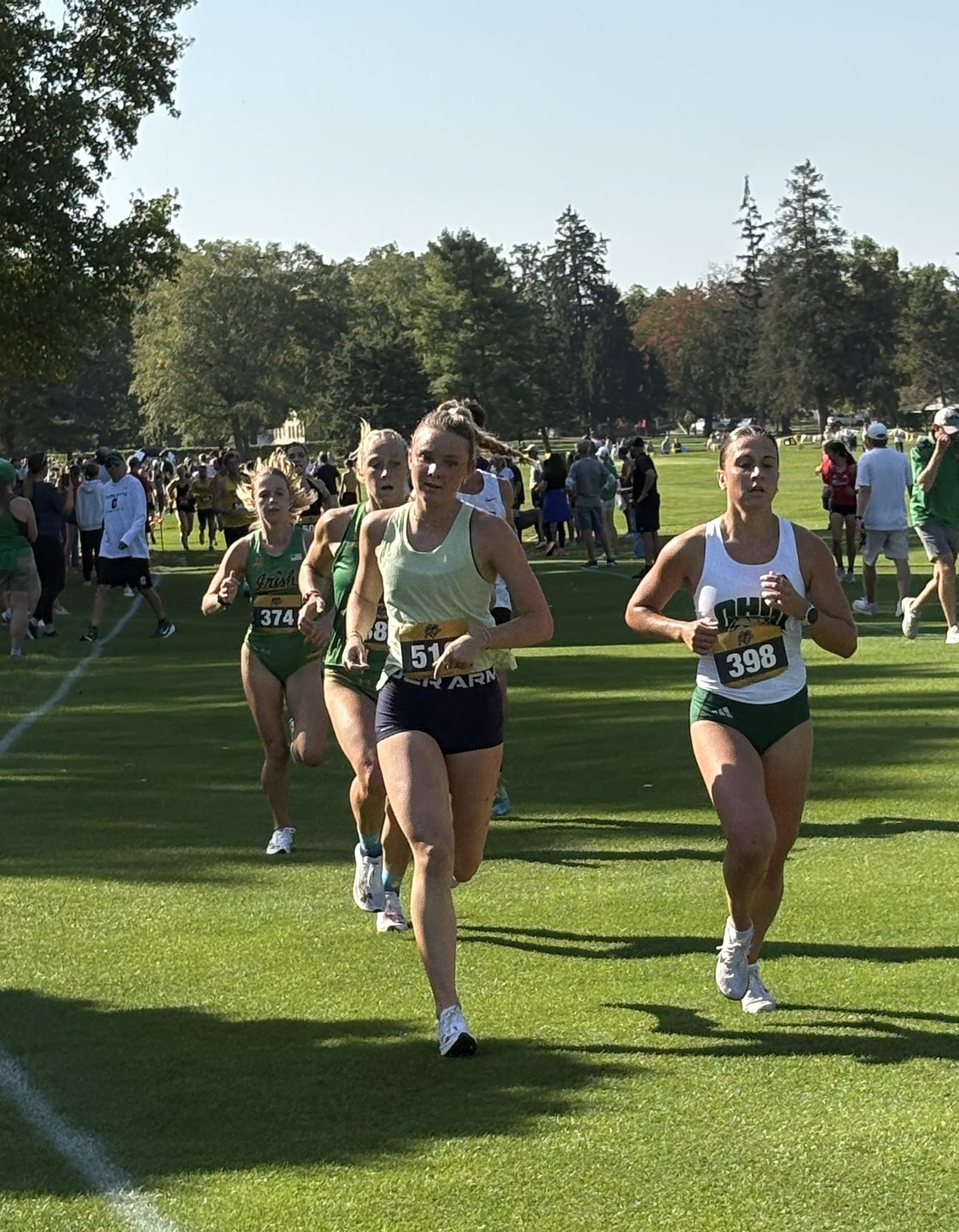 Female runners participating in a cross-country race on a grassy field with trees and spectators in the background.