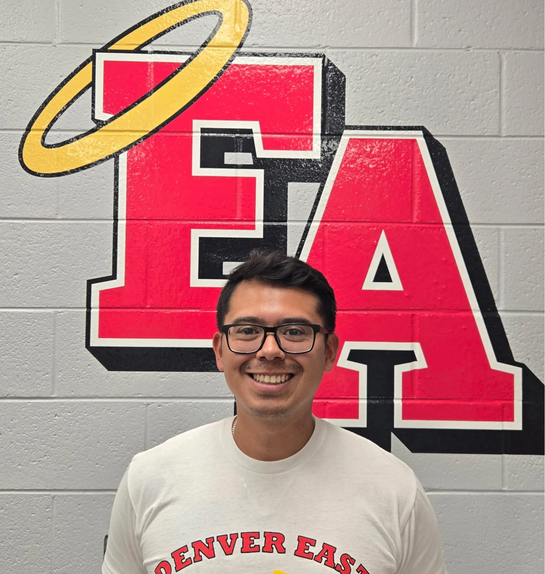 A smiling man with glasses and a white Denver East t-shirt standing in front of a gray brick wall with a large red and black logo that says 'EASTER' topped with a yellow halo and some yellow ring.