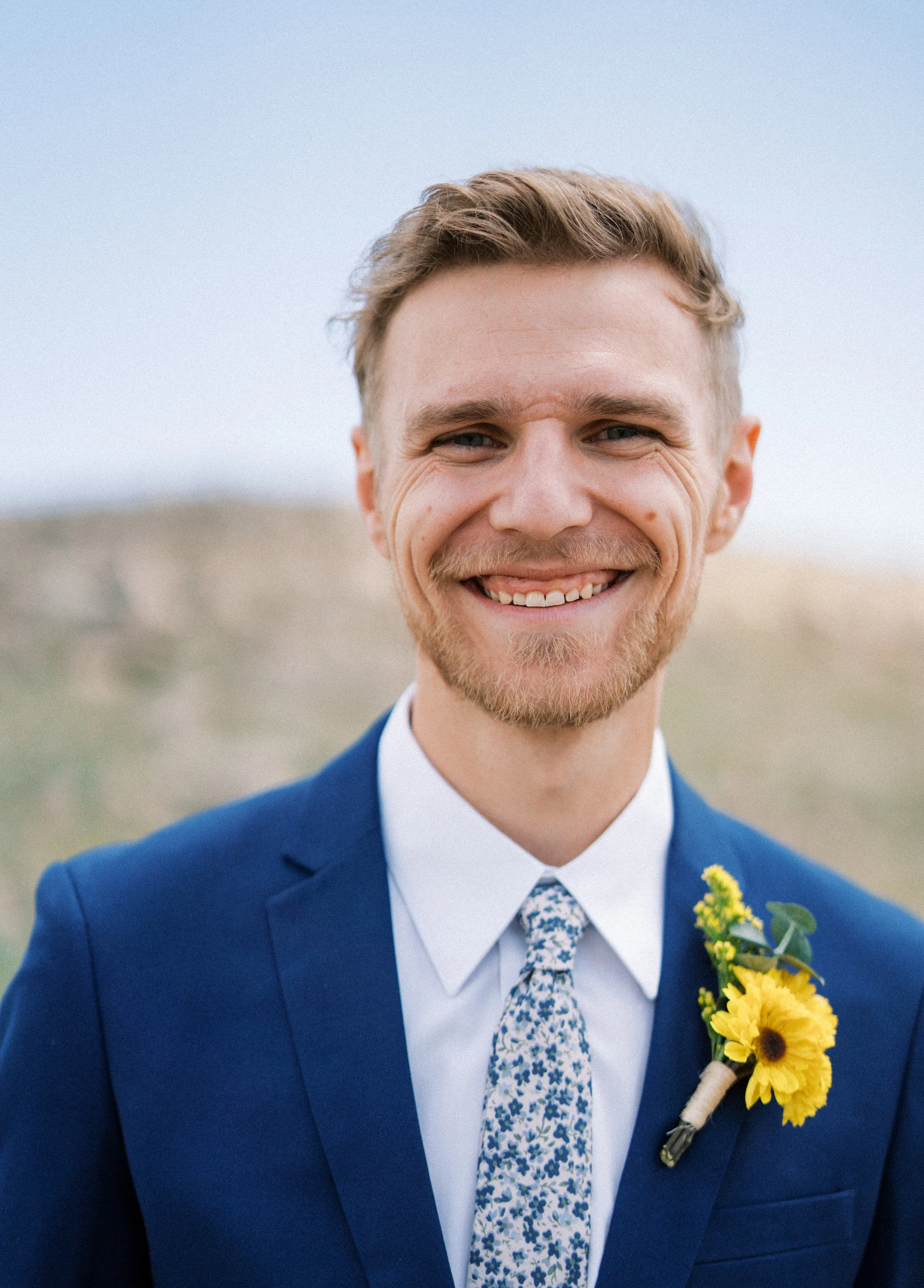 A smiling man wearing a blue suit, white shirt, and floral tie, with a yellow floral boutonniere, standing outdoors against a blurred background.