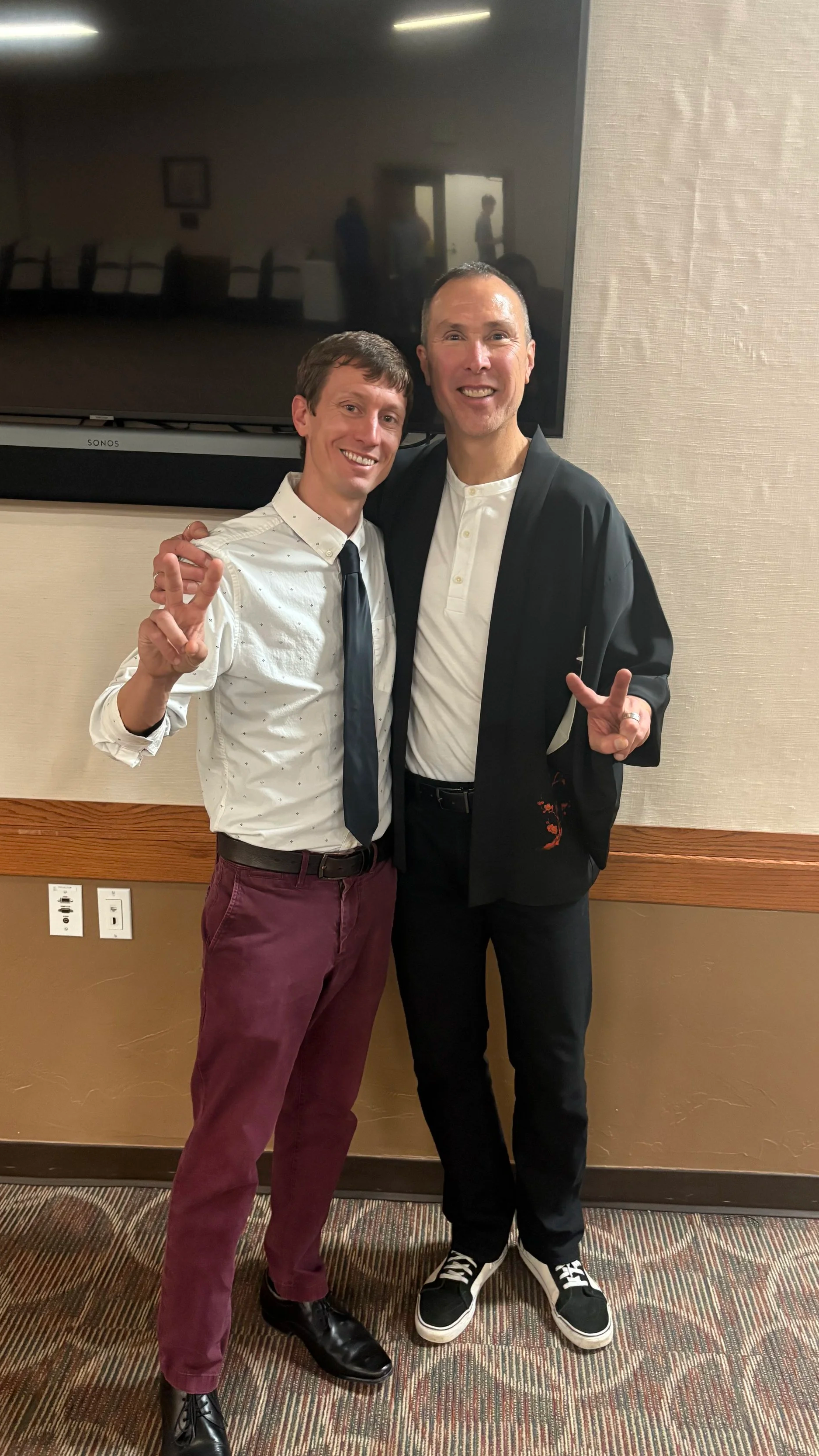 Two men smiling and posing together indoors, one dressed in a white shirt, tie, and maroon pants, the other in a black jacket, white shirt, and black pants.