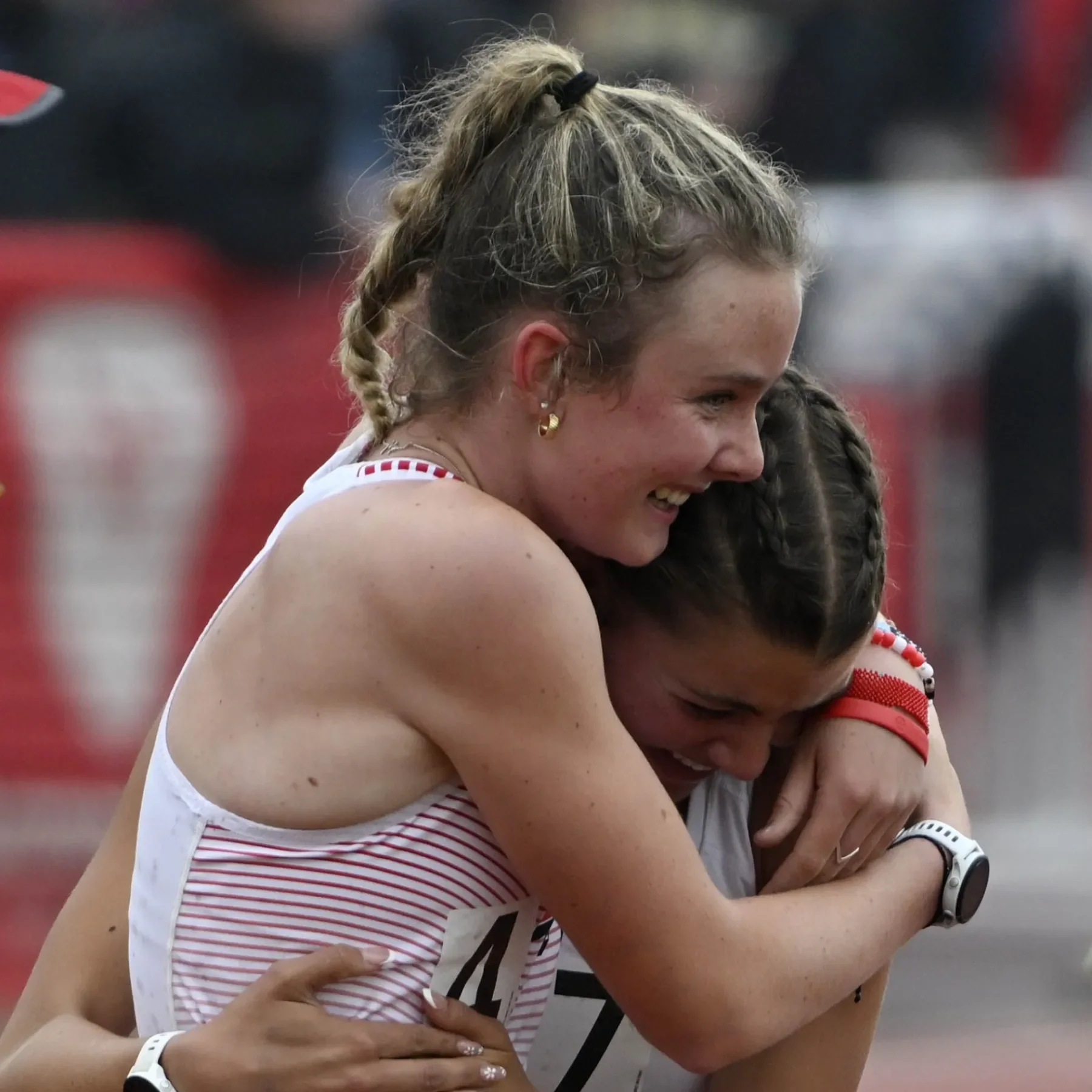 Two women hugging each other, smiling, after a race. One woman is wearing a red and white athletic outfit and the other is wearing a gray shirt. They appear happy and emotional.