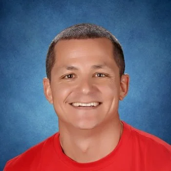 A young man with short brown hair and a big smile, wearing a red shirt, against a blue background.