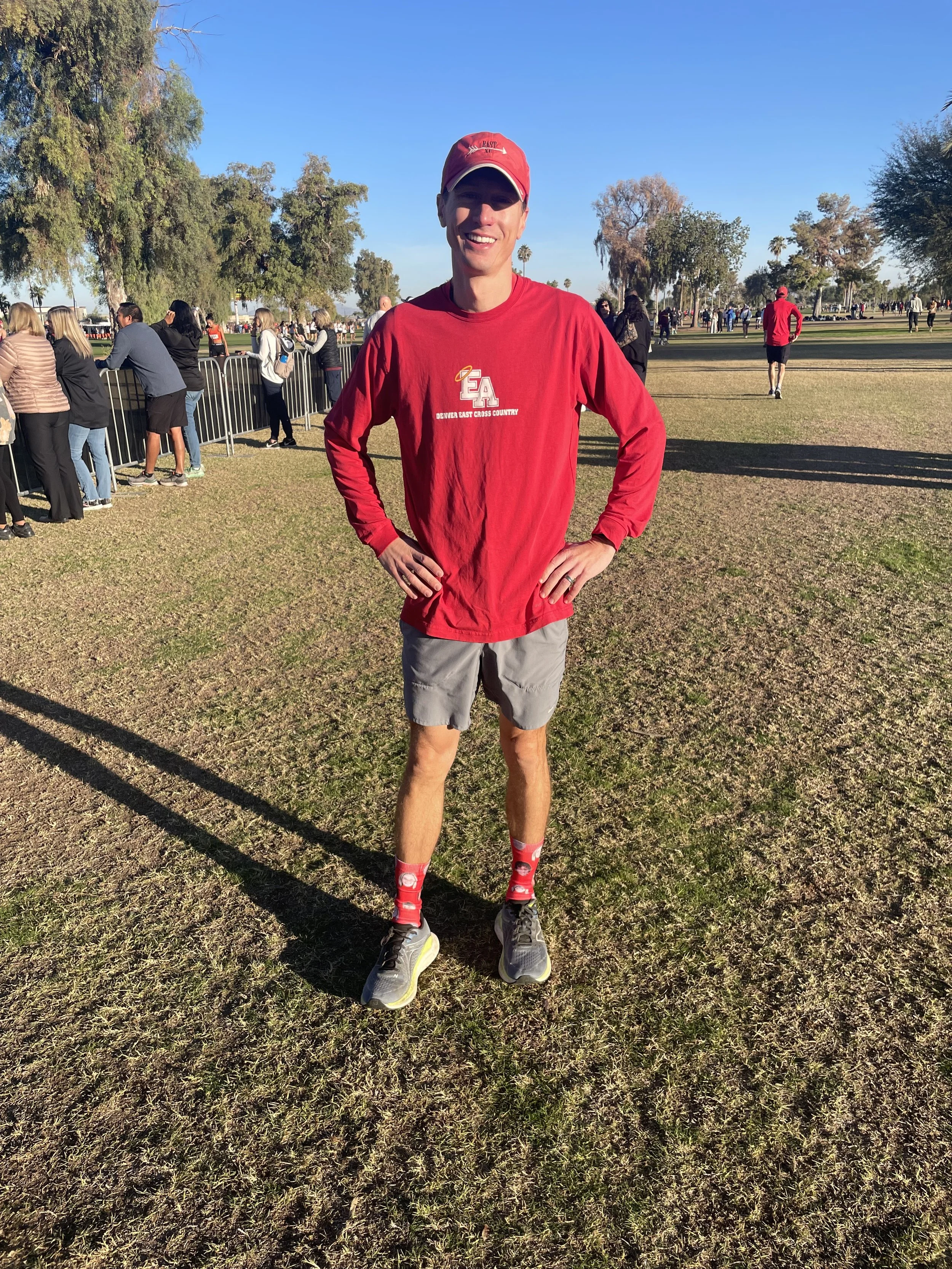A young man standing outdoors on a grassy field, wearing a red long-sleeve shirt, gray shorts, red patterned socks, and gray running shoes, smiling at the camera with hands on his hips. There are trees and a crowd of people behind him, with some standing by a metal fence, during a sunny day.
