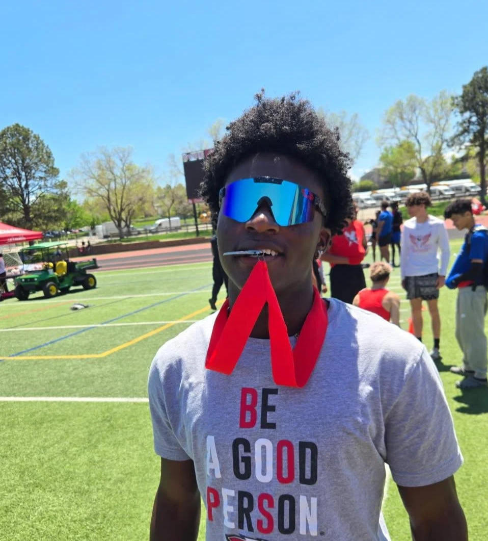 A young person wearing blue reflective sunglasses and a gray T-shirt with the words 'BE A GOOD PERSON' in red, black, and white, standing on a sports field during a sunny day. They have a red ribbon around their neck and a lollipop in their mouth.