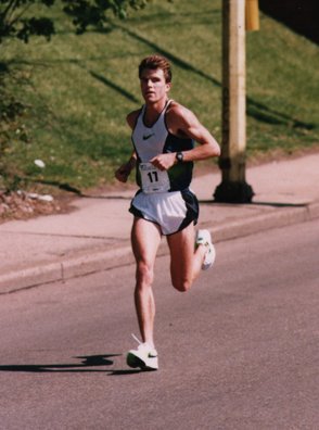 A young male runner wearing a white tank top, gray shorts, and white running shoes running on a paved road with grass and a wooden utility pole in the background.