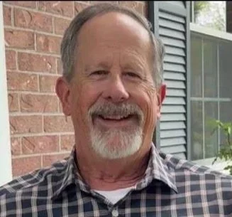 A smiling middle-aged man with a beard and mustache standing outside in front of a brick wall and window with shutters.