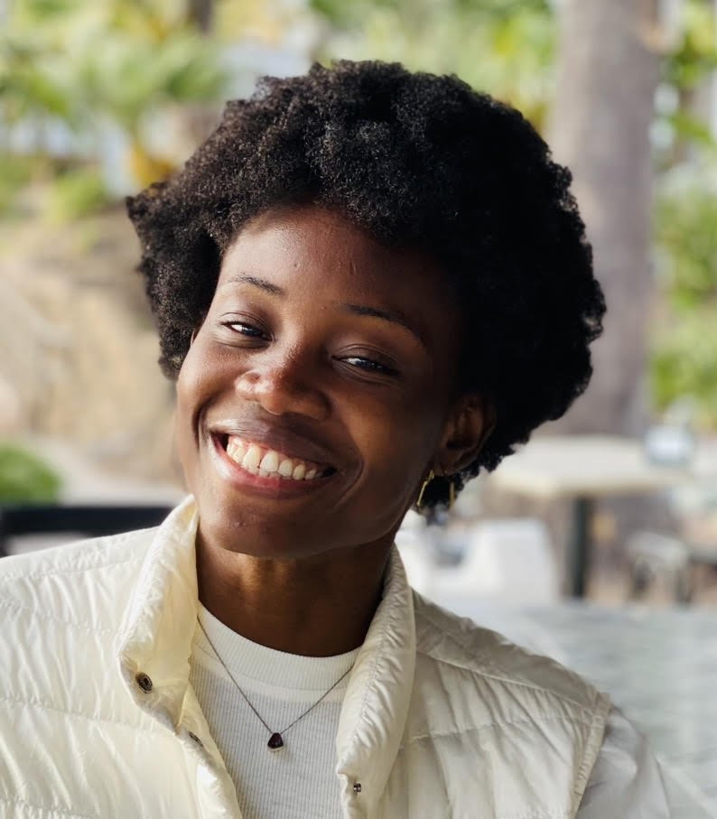 Portrait of a smiling black woman with short curly hair, wearing a cream-colored vest and a necklace, outdoors with blurred green trees and buildings in the background.