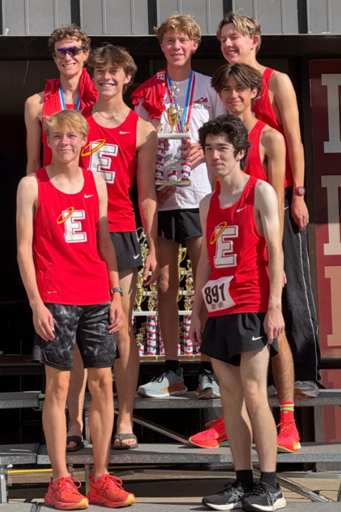 Seven young athletes in red and white uniforms standing on a podium, celebrating with medals and trophies after a race.