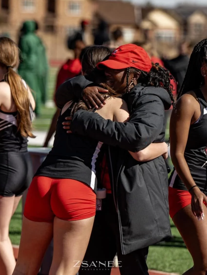 A woman in a black and red athletic outfit hugging a smiling coach wearing a red cap and black jacket during a sports event on a track field.