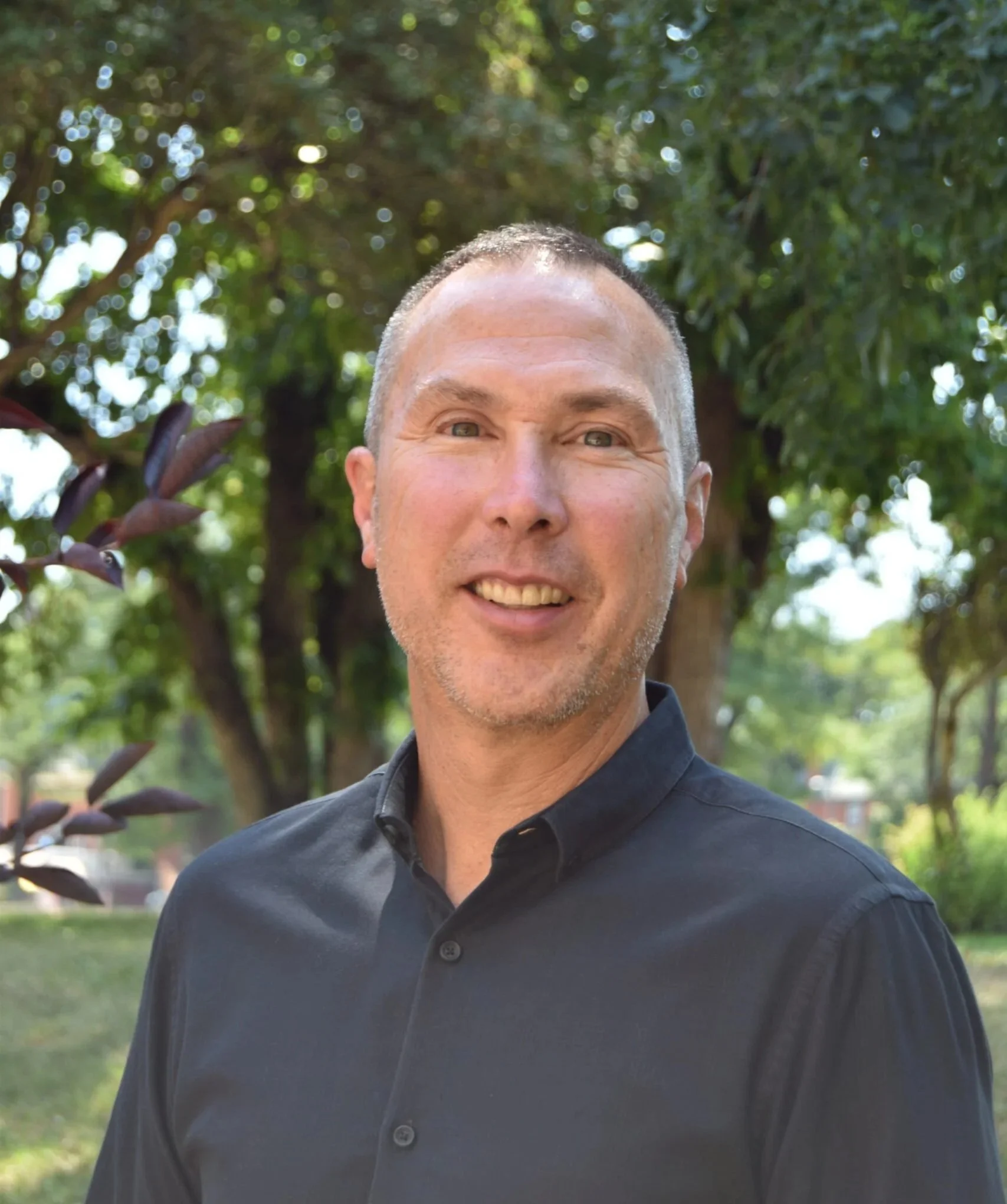A smiling man with short hair and a trimmed beard, wearing a black collared shirt, standing outdoors with trees and greenery in the background.