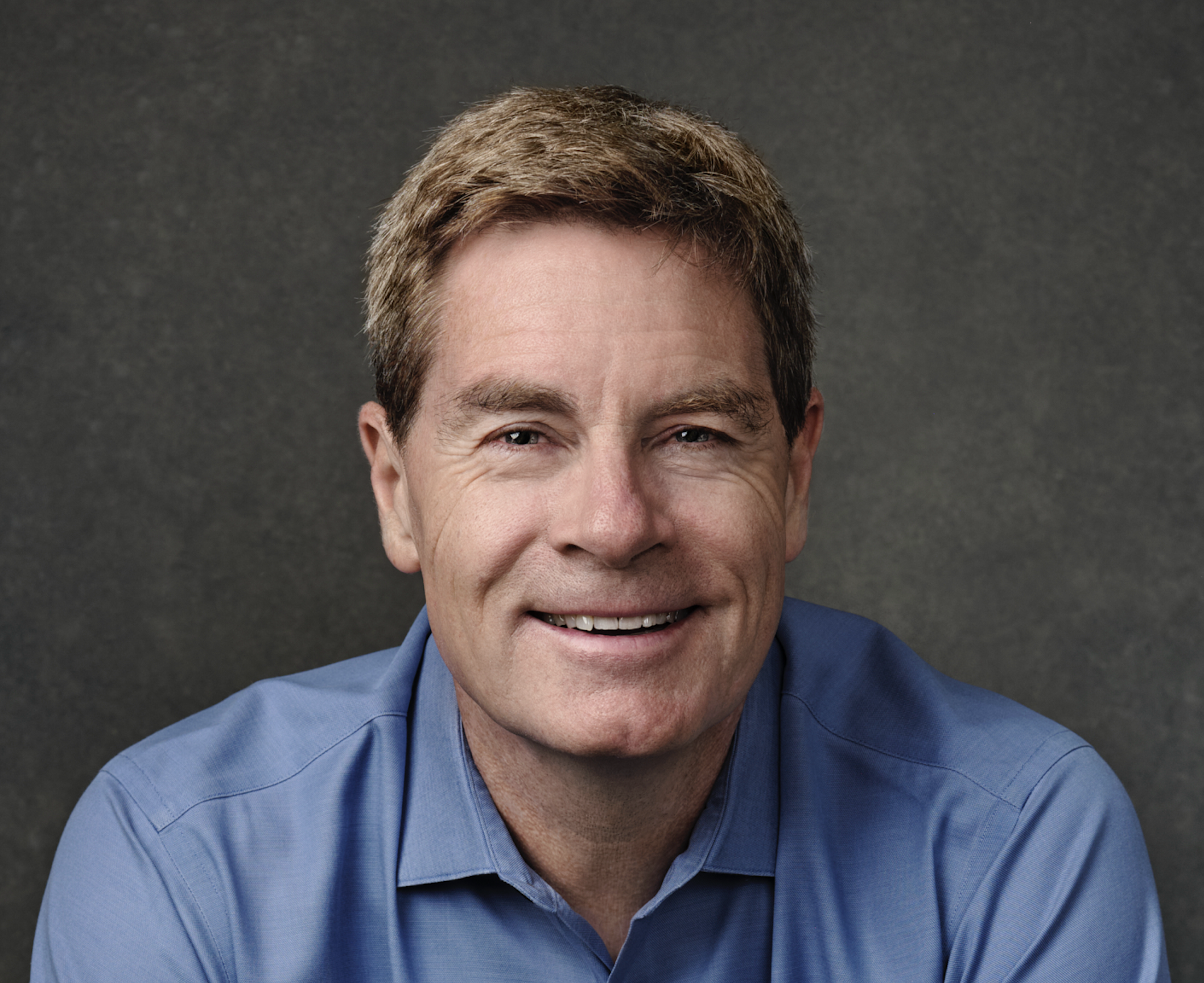 A middle-aged man with light skin, brown hair, and blue eyes wearing a blue collared shirt, smiling at the camera against a dark gray background.