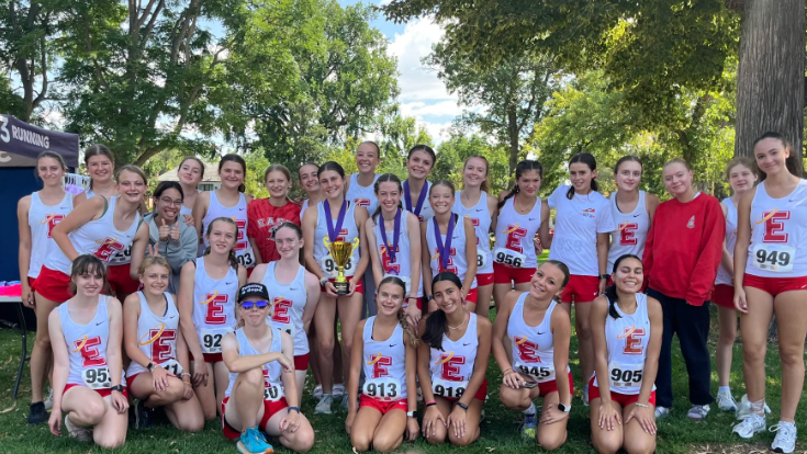Group of young female athletes in track uniforms posing outdoors after a race, some holding medals and a trophy, surrounded by trees.