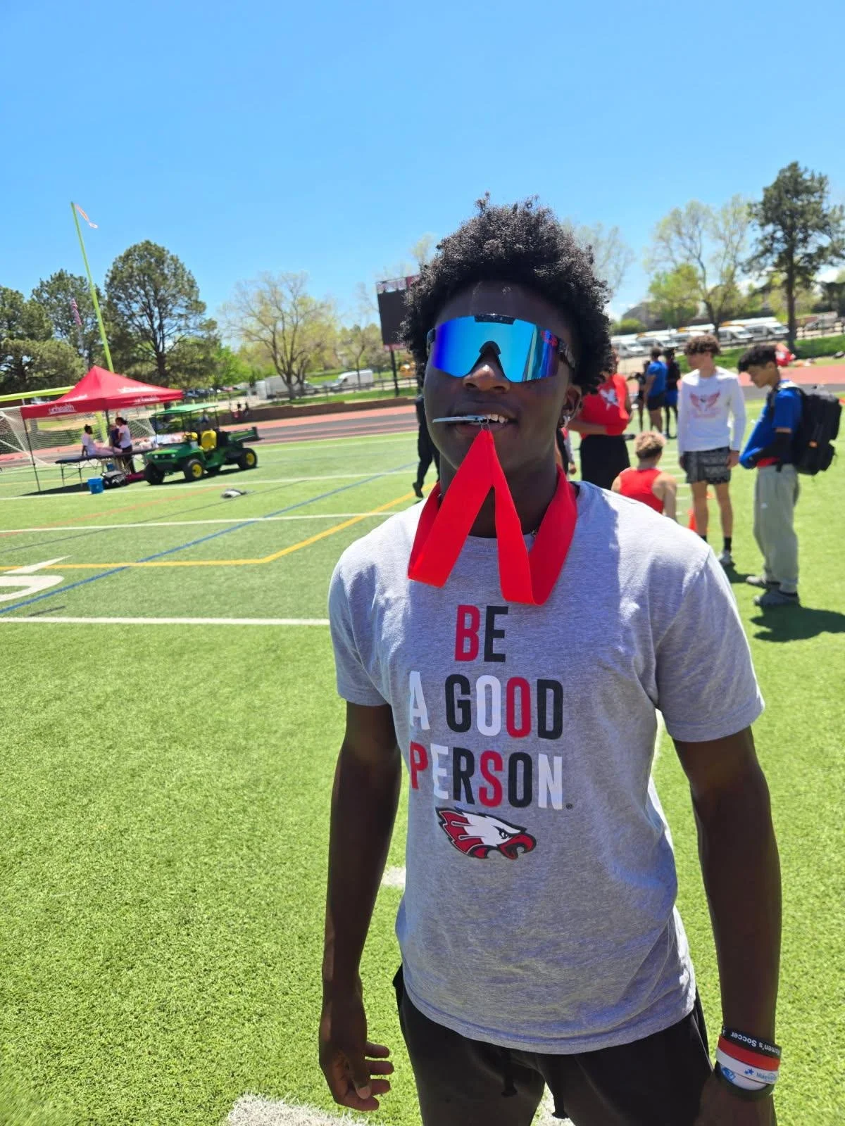 Young man in athletic gear with sunglasses and a medal around his neck, standing on a sports field on a sunny day.