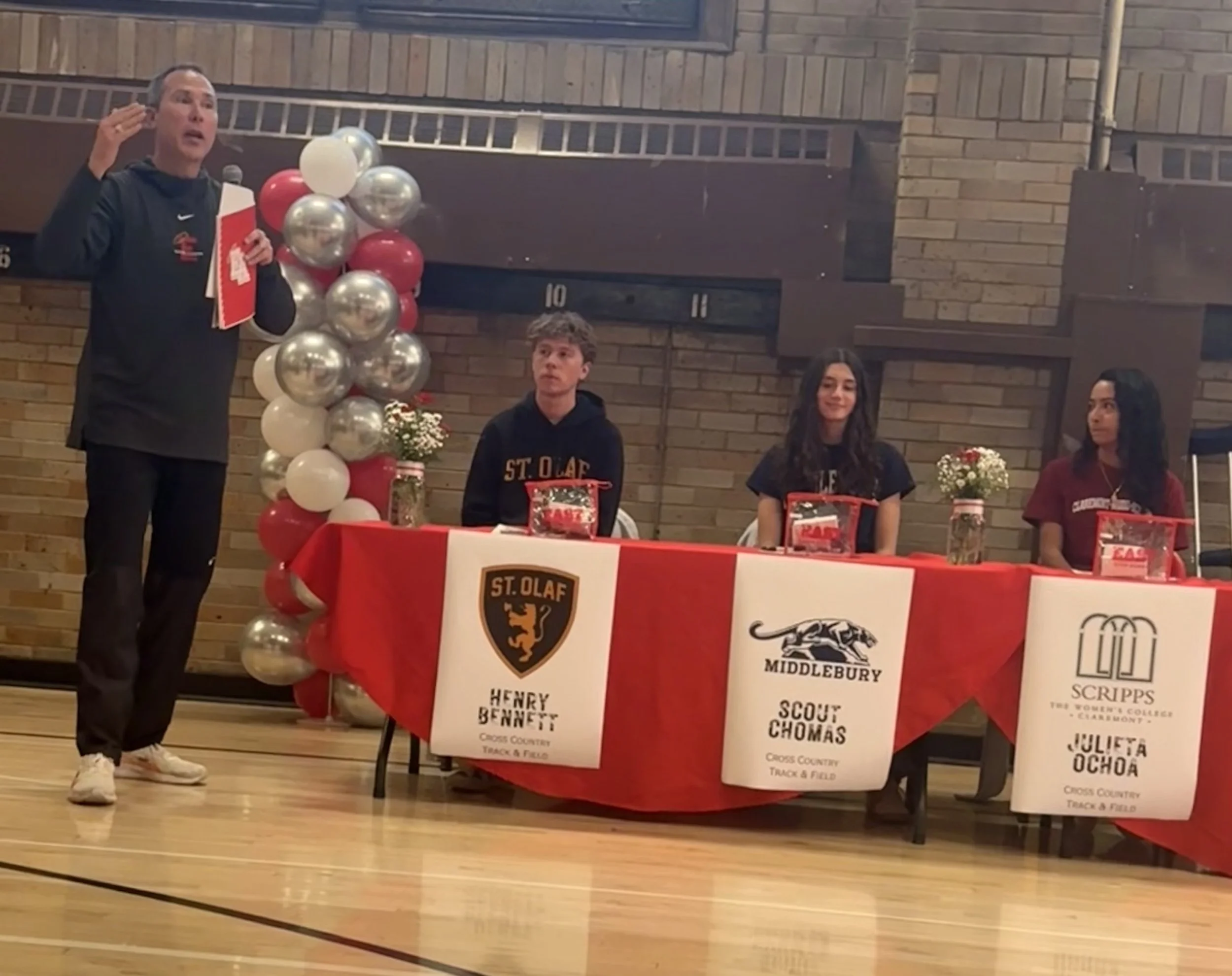 A man is giving a speech at a table with three young women behind it, decorated with balloons and flowers, in a gymnasium. The table displays signs with the names and logos of cross country teams, including St. Olaf, Middlebury, and Scripps College.