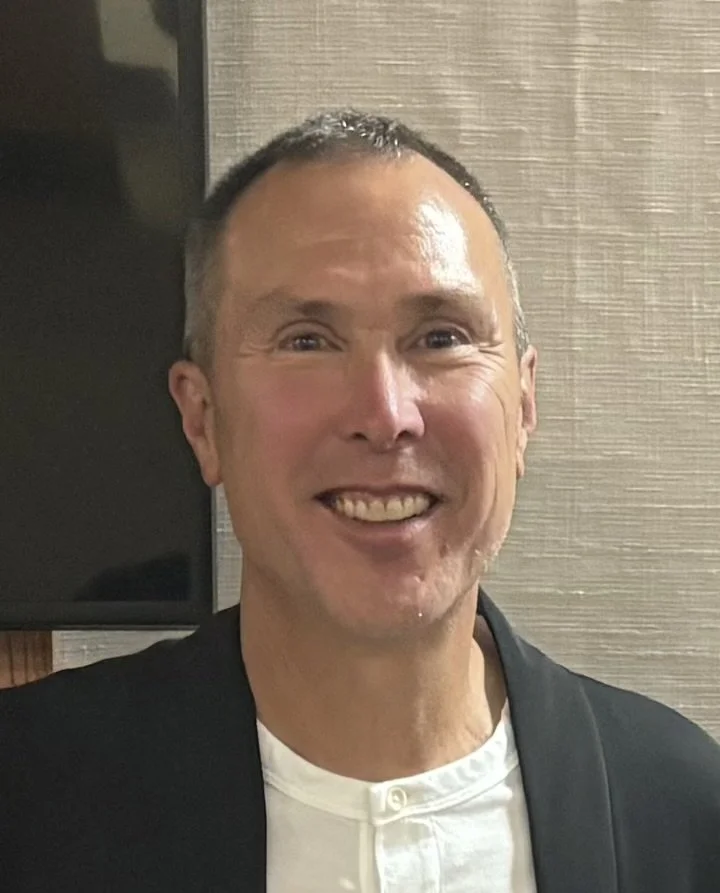 A smiling man with short hair and wearing a black blazer over a white shirt standing indoors against a textured wall.