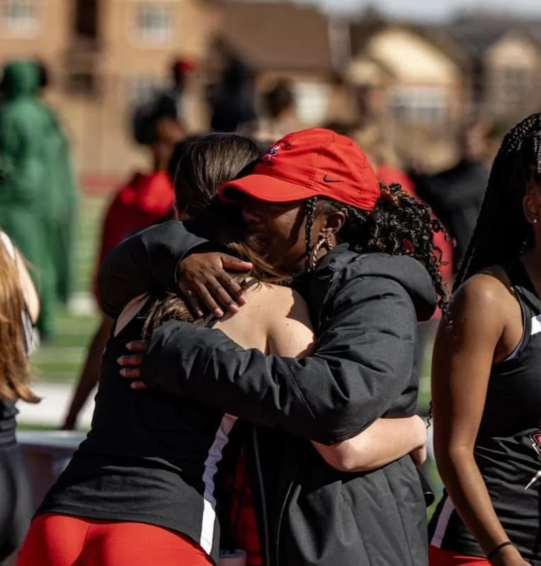 Two women hugging at an outdoor sports event, with other people and buildings in the background.