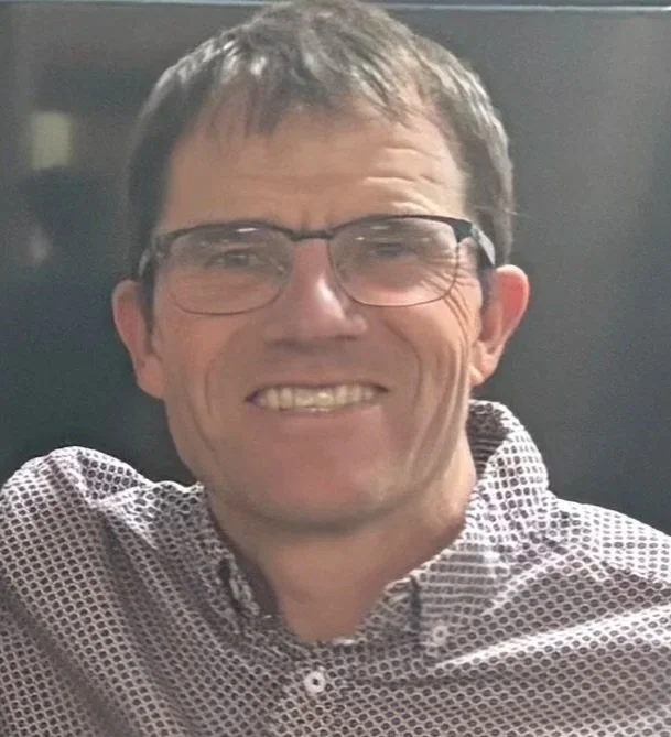 Smiling middle-aged man with glasses and short brown hair, wearing a patterned button-up shirt.