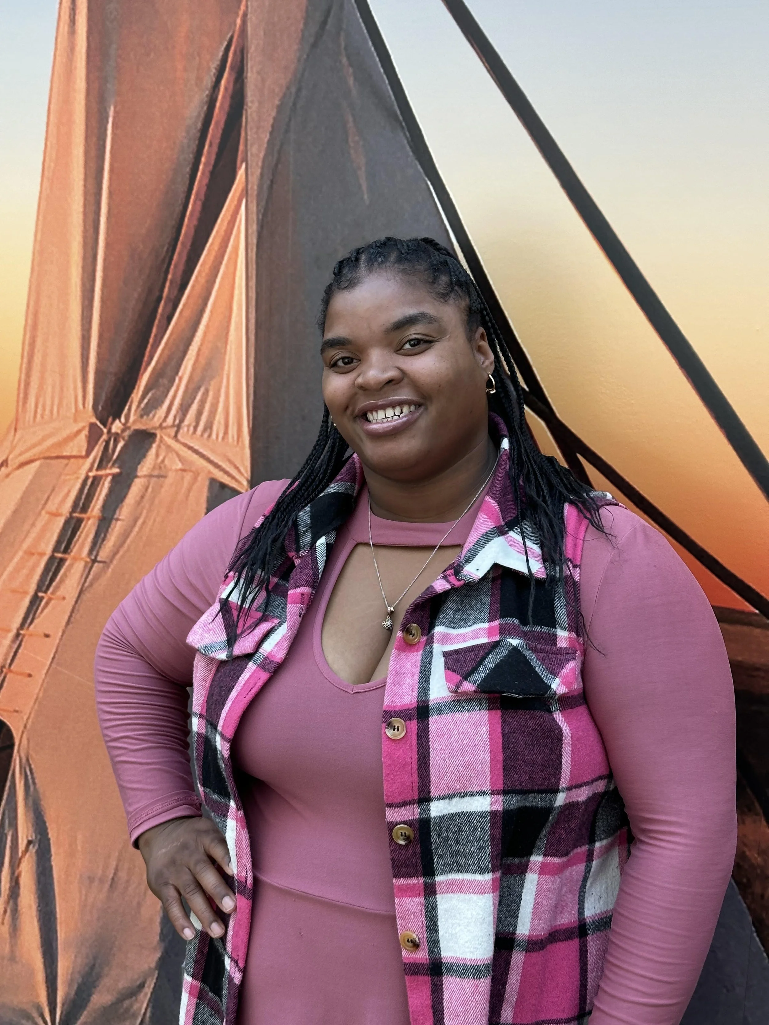 A woman in a pink long-sleeve shirt and plaid vest standing in front of a large orange and black tent backdrop, smiling at the camera.
