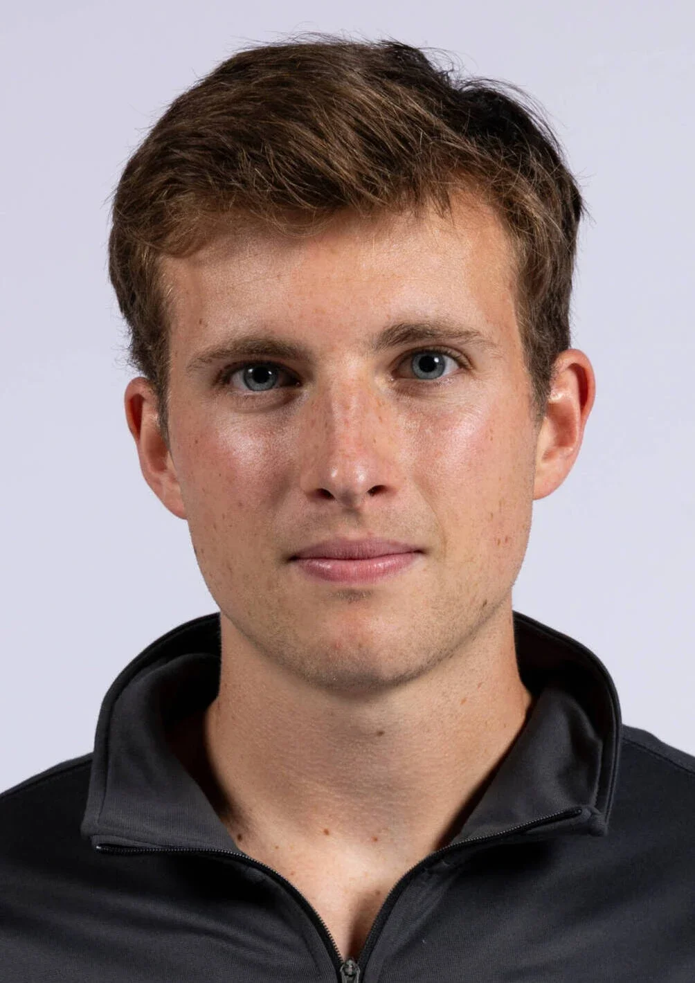 A young man with short brown hair, blue eyes, and fair skin, wearing a black zip-up jacket, looking directly at the camera against a plain light background.