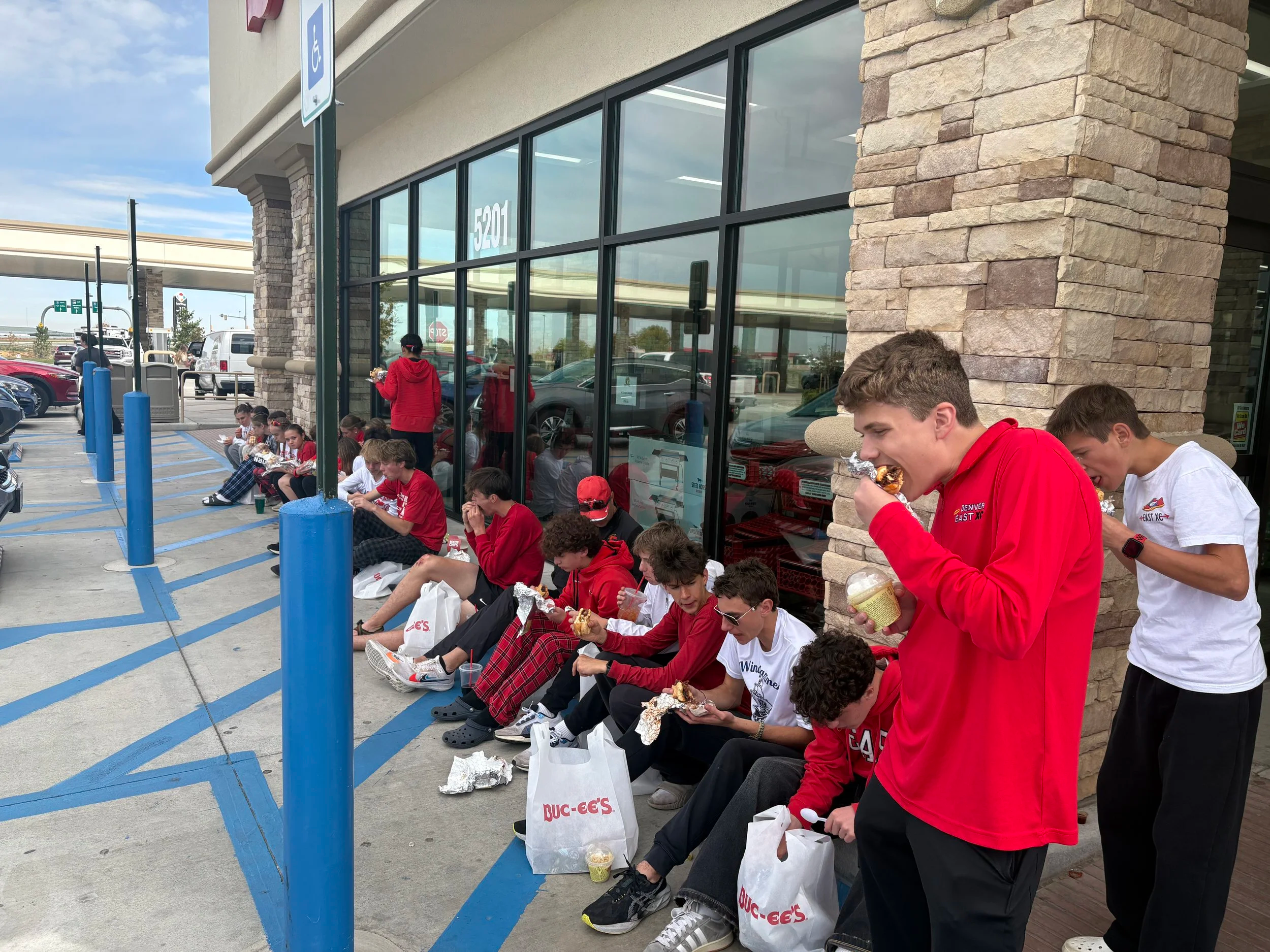Group of kids and teenagers sitting and standing outside a fast-food restaurant, eating. Some are sitting on the sidewalk while others stand. They are wearing casual clothes, including red and white shirts. There are take-out bags from BUC-EES, parked cars, and a handicap parking sign visible.
