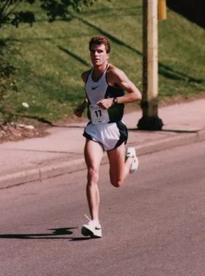 A young male runner in a white tank top and shorts running on a paved road during the day.