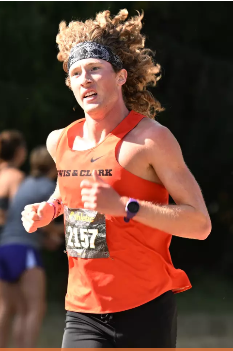 A female runner wearing an orange tank top with 'Lewis & Clark' and a race bib number 2457, running outdoors with other participants in the background.