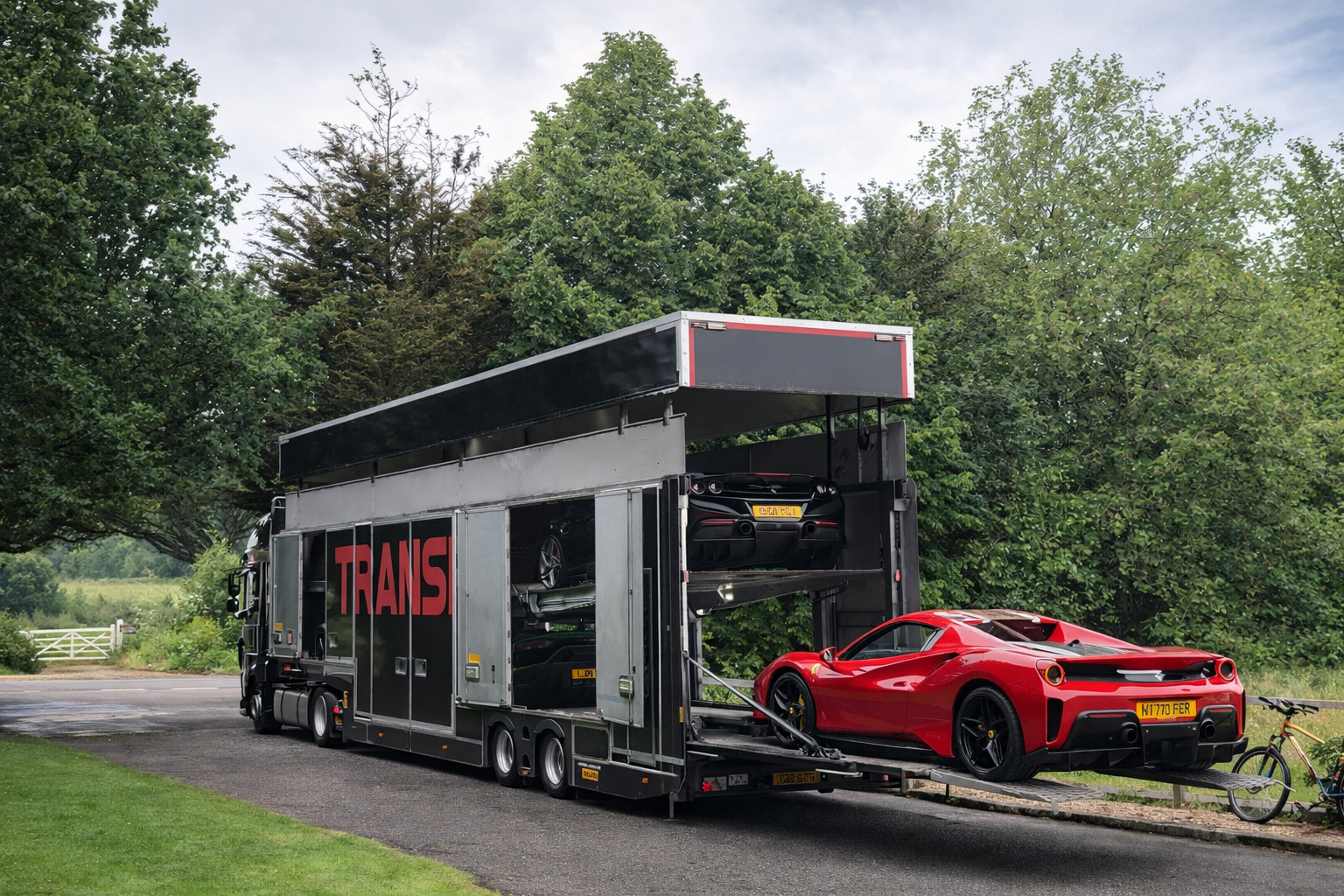 A car transport truck carrying two sports cars, one red and one black, parked on a country road surrounded by green trees.