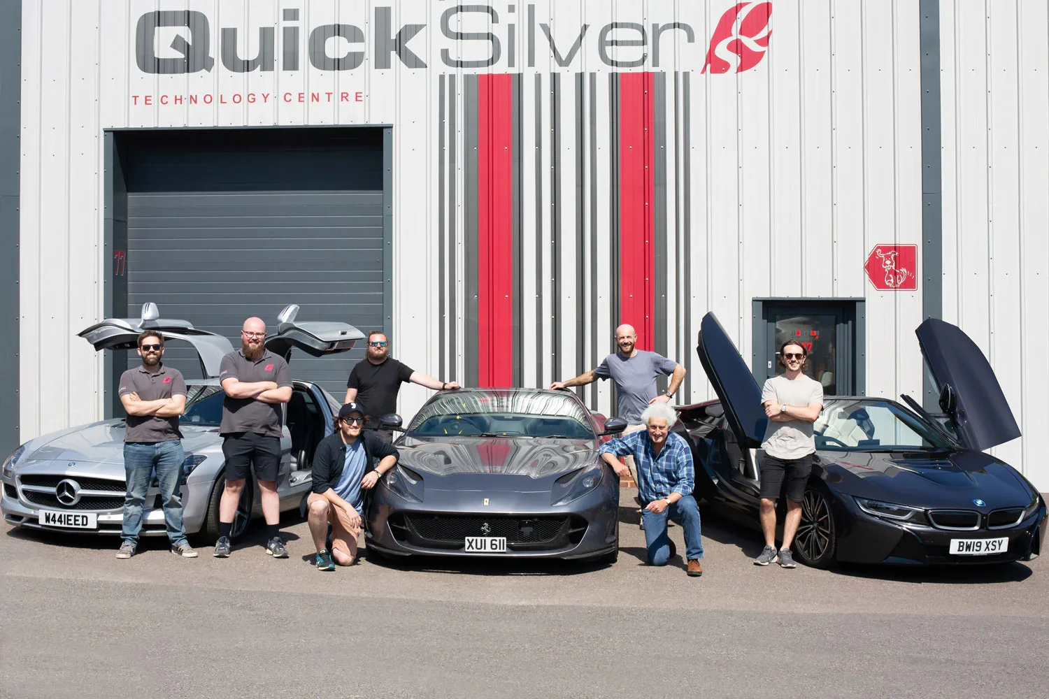 Group of seven men posing with luxury cars in front of the Quicksilver Technology Centre building. There are three sports cars: a silver Mercedes-Benz, a black Ferrari, and a black BMW i8 with their doors open or raised.