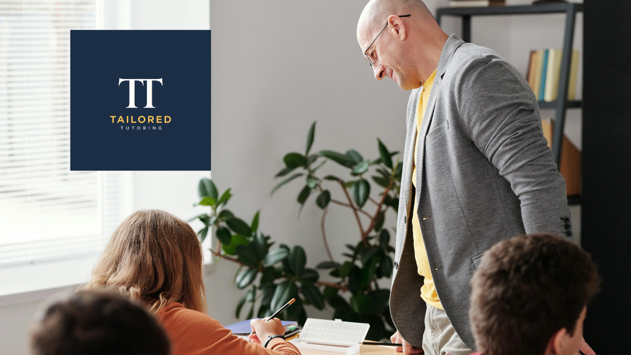 A tutor is standing and leaning over a desk, engaging with students in a classroom or tutoring setting. The students are seated at the desk, and one of them is taking notes. There is a large plant in the background and window blinds letting in natural light. The logo of 'Tailored Tutoring' is present in the upper left corner.