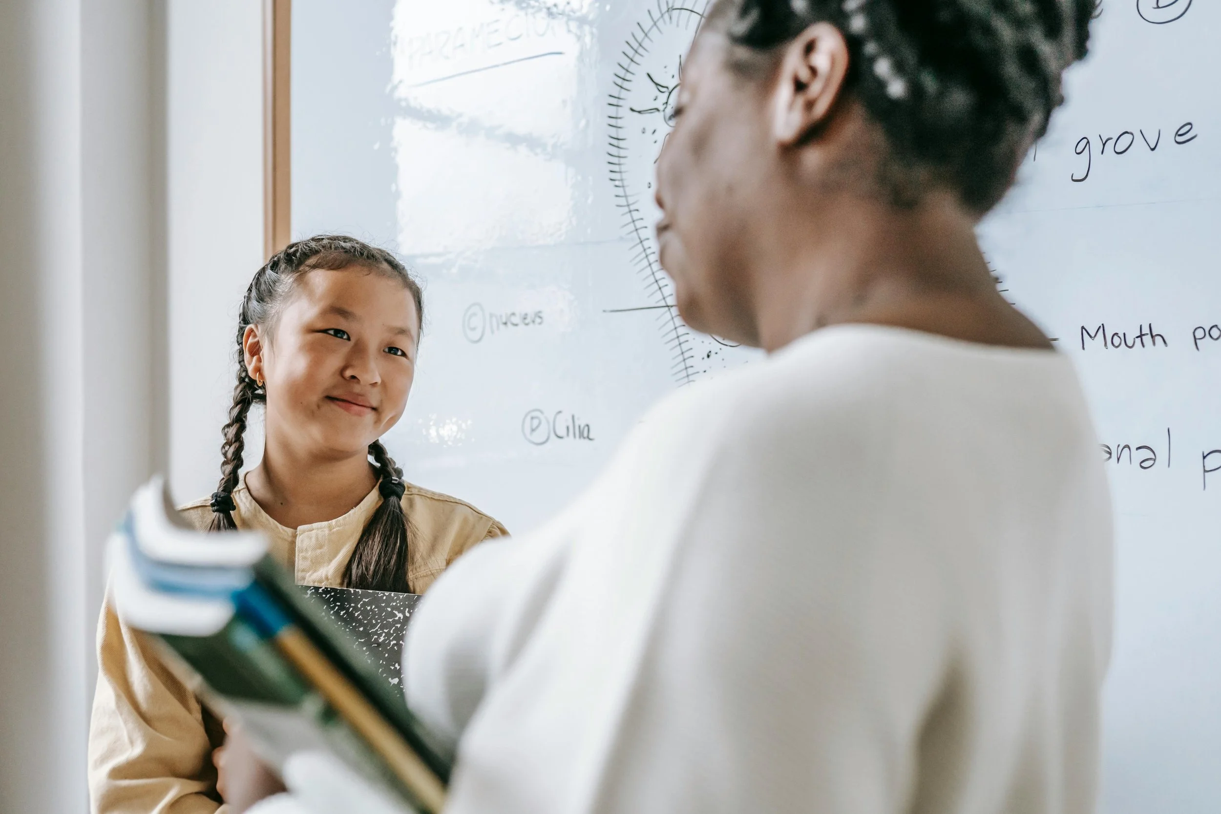 A young girl with braided hair smiling and looking at an adult woman in a classroom. The woman is holding a notebook and a pen, facing a whiteboard with handwritten notes and drawings.