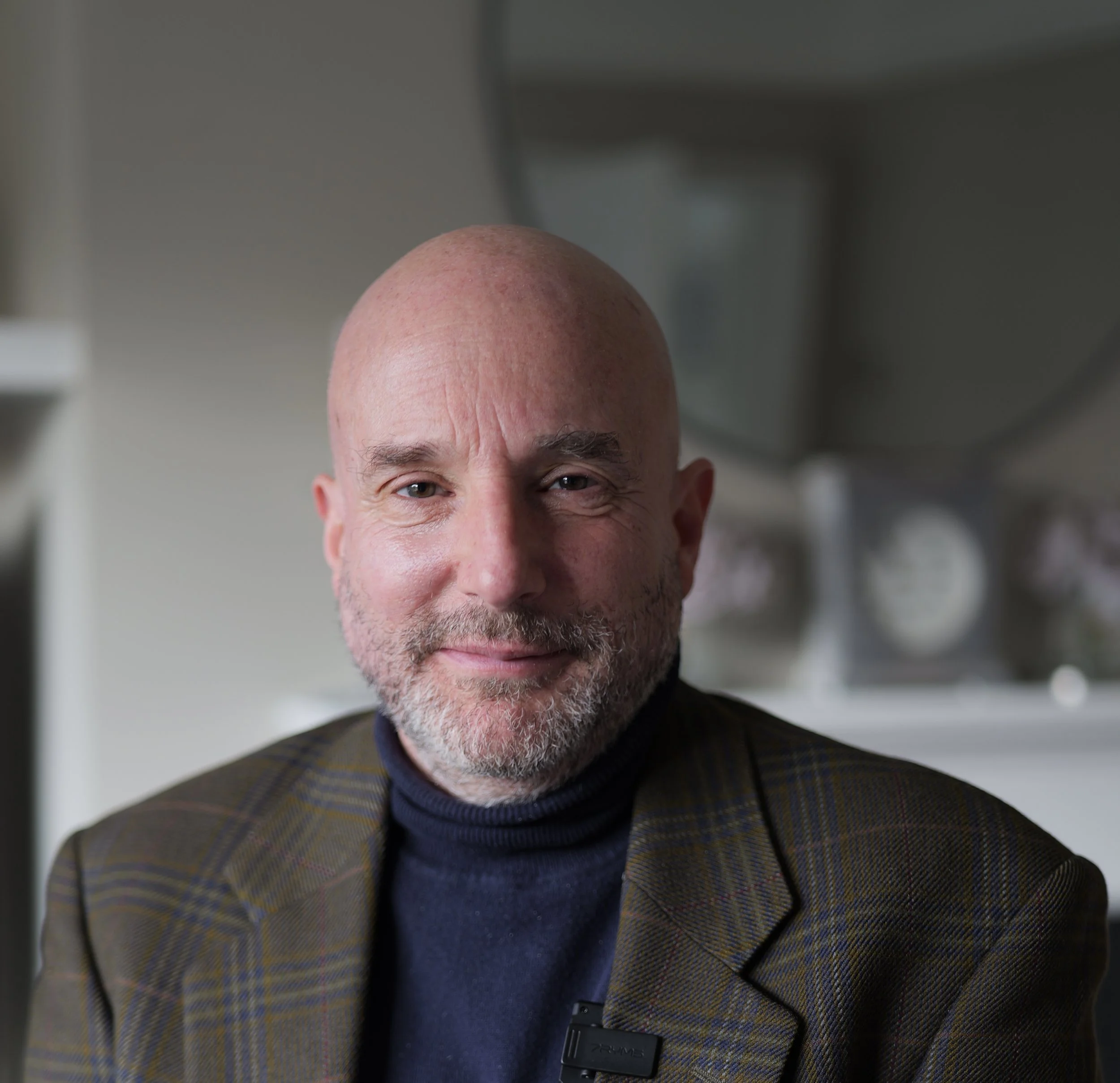 Close-up of smiling bald man with beard, wearing a dark turtleneck and a plaid blazer, sitting indoors.
