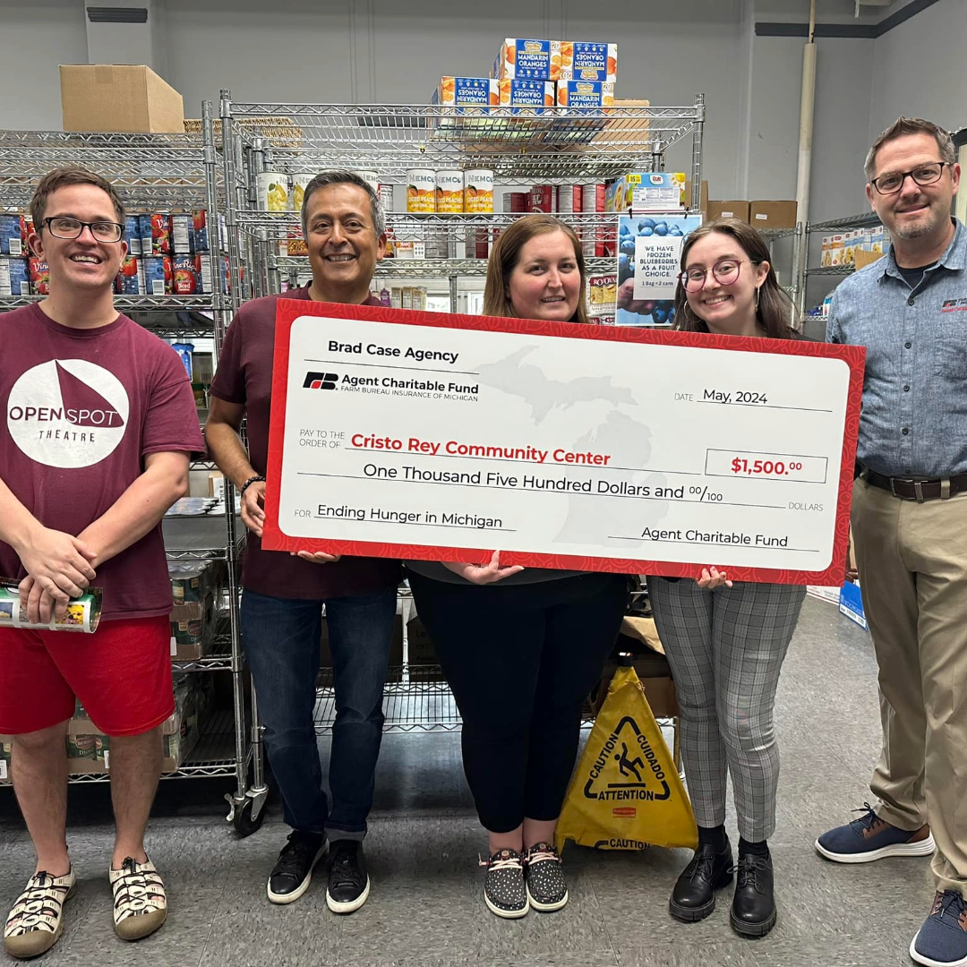 Group of five people holding oversized check for $1500, standing in a storage room with shelves of canned and boxed food, celebrating donation to Cristo Rey Community Center.