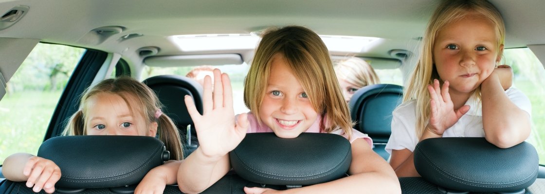 Three young girls sitting in the backseat of a car, smiling and waving at the camera.