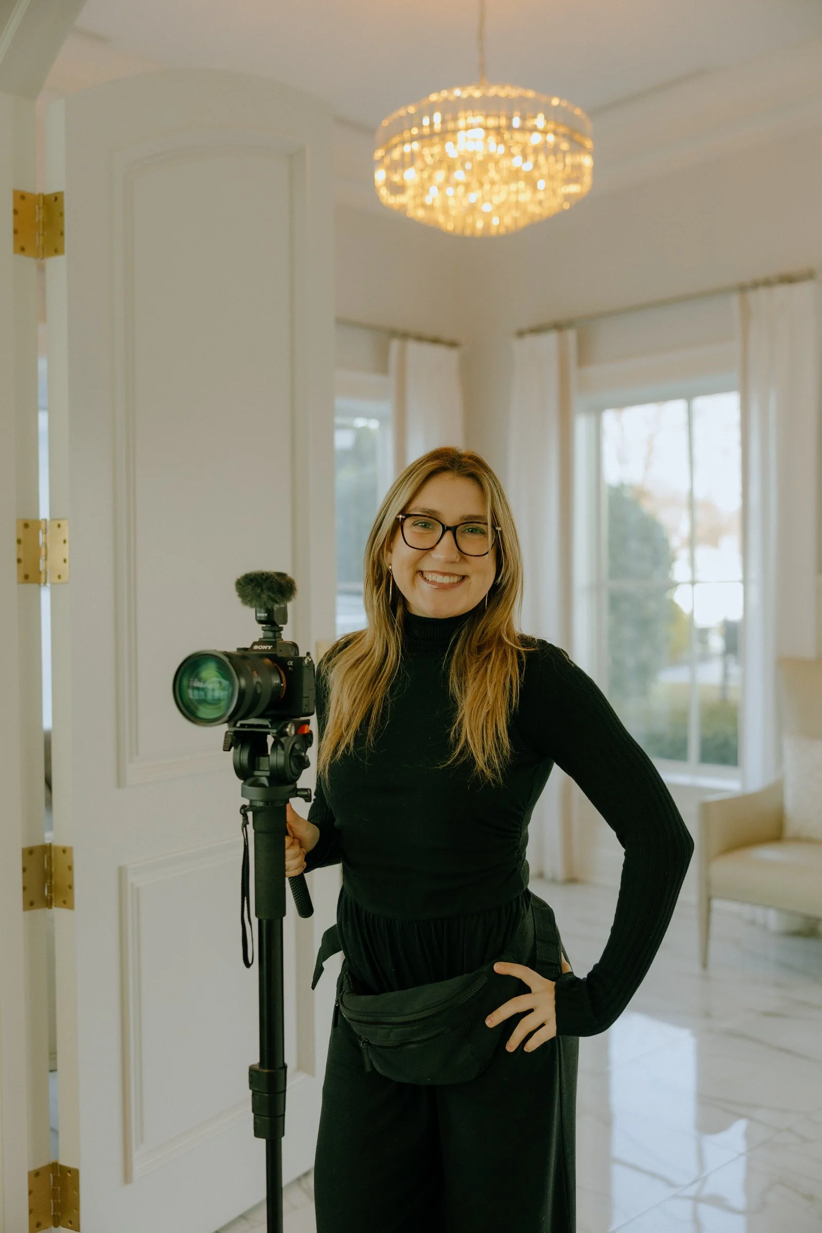 A young woman with glasses, long hair, and a smile, standing indoors with a camera on a tripod, in a well-lit, elegant room with large windows, white curtains, and a chandelier.