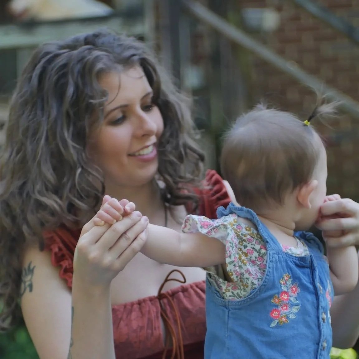 A woman with curly brown hair smiling while holding a young girl's hand outside near a metal and brick structure.