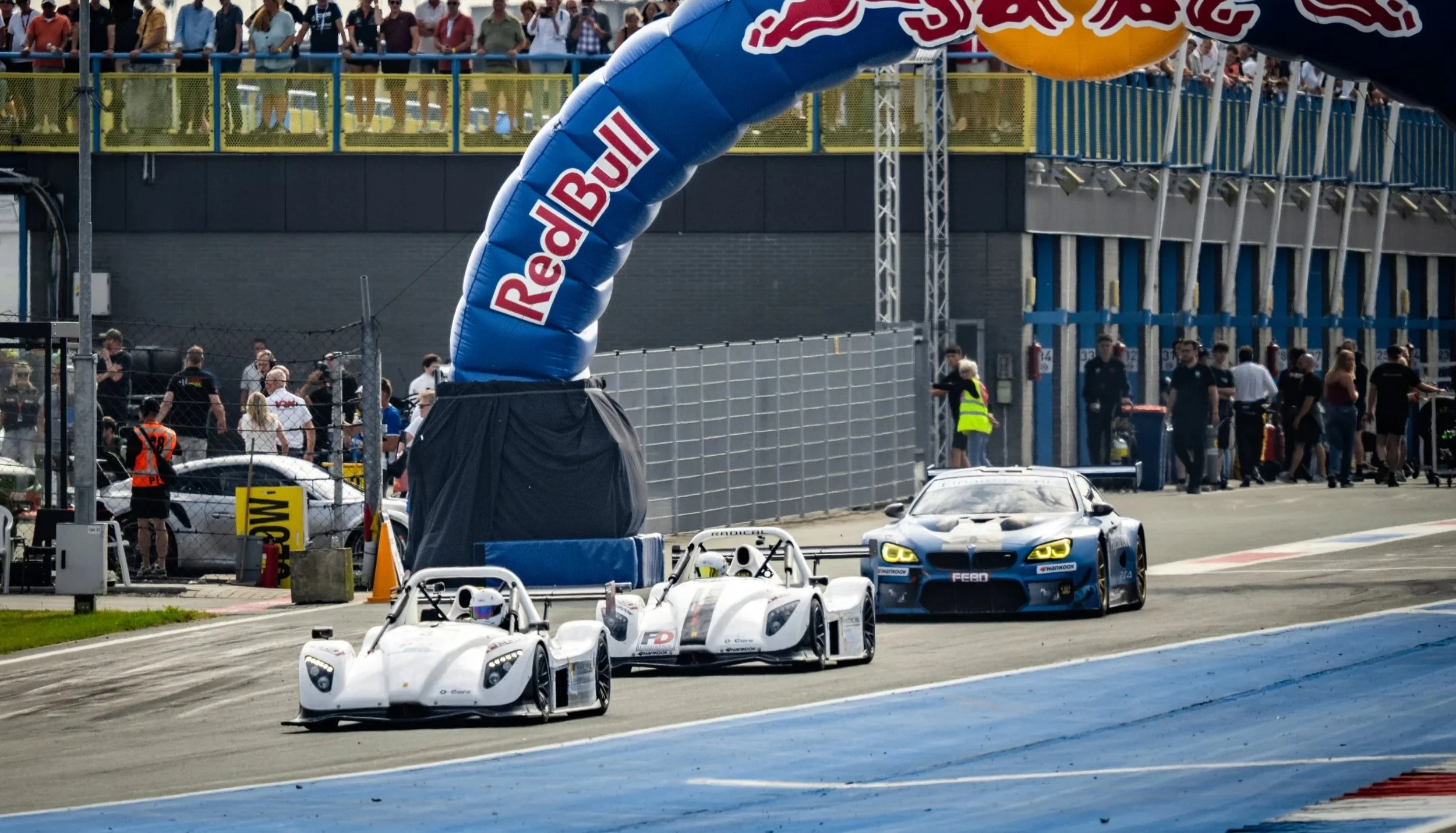 Race cars on the track during a racing event, with spectators watching from the stands and a large inflatable Red Bull arch over the finish line.