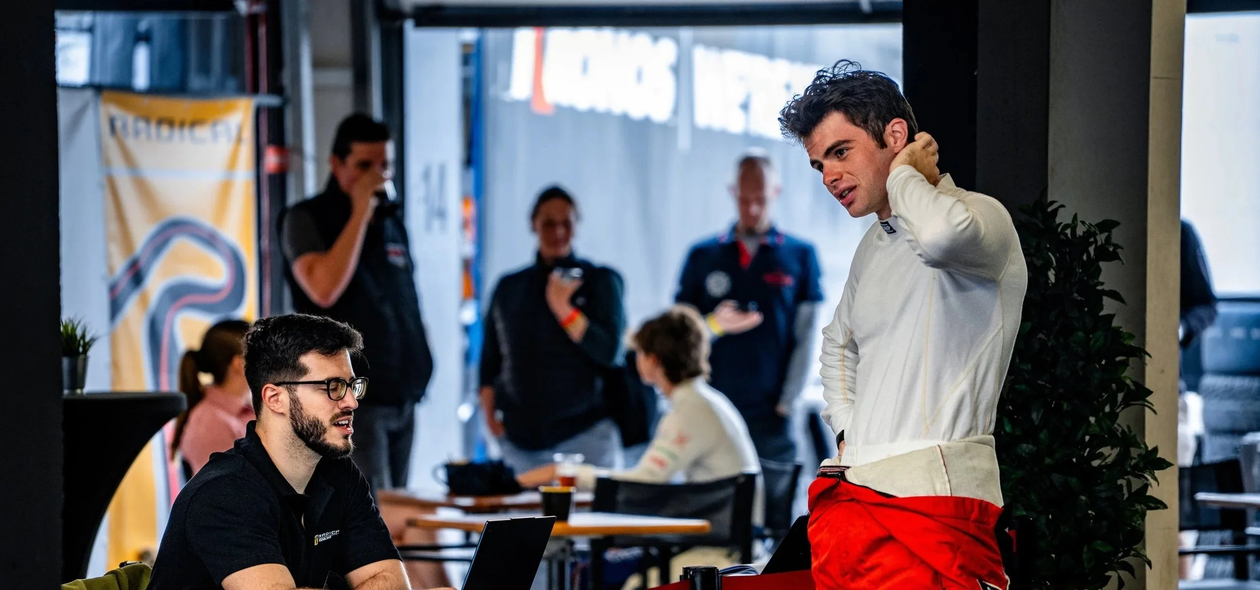 Two men are in a garage or workshop, with one man sitting at a table with a laptop and the other standing, dressed in racing gear. Several people in racing uniforms are in the background, some using their phones, and there are banners and plants arou