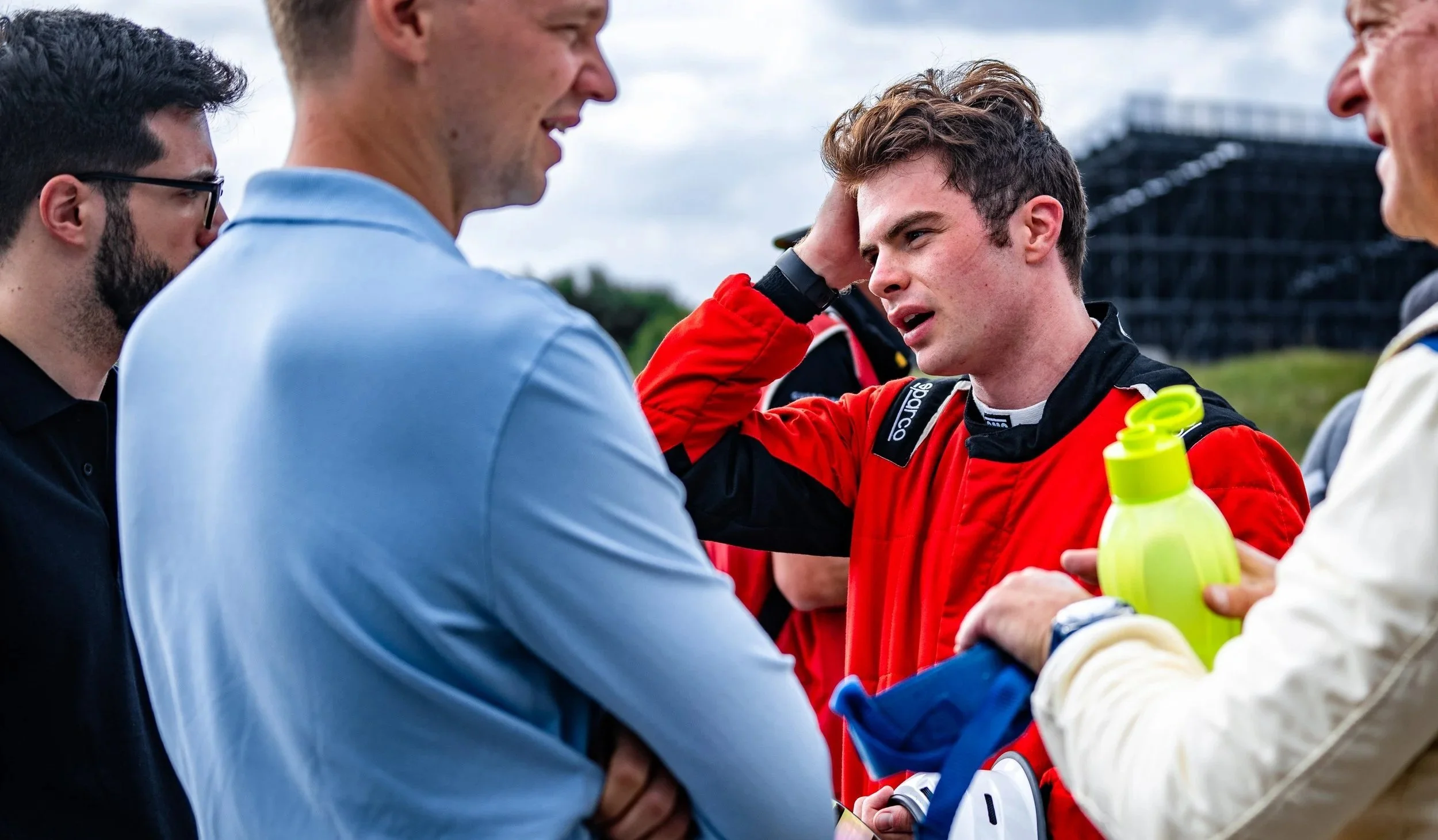 Driver Miles Wragg in a red suit talking with a group of men at a racing event.