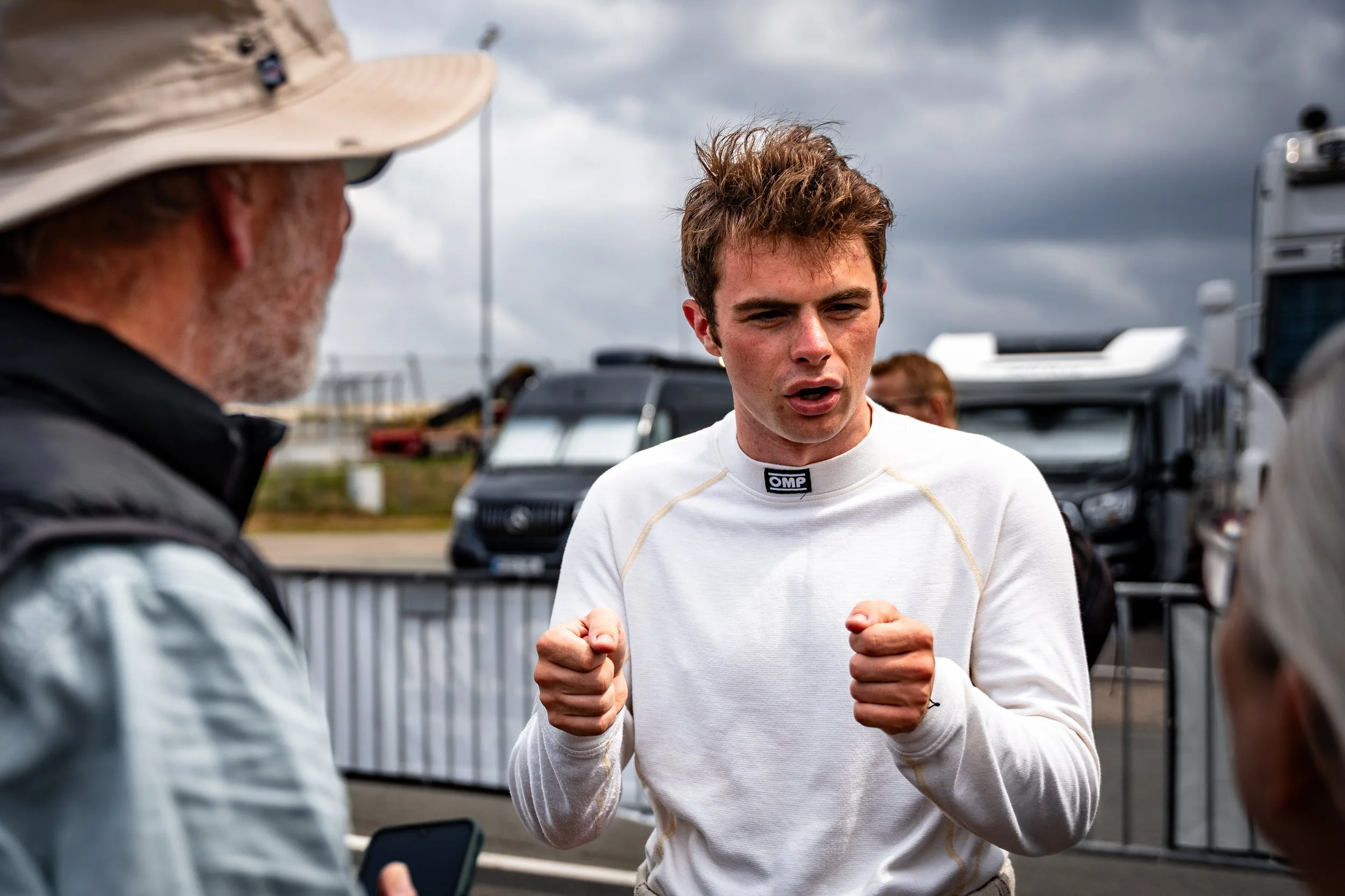 Miles Wragg in white racing suit talking to older man in sunglasses and hat at a race track outside on a cloudy day.