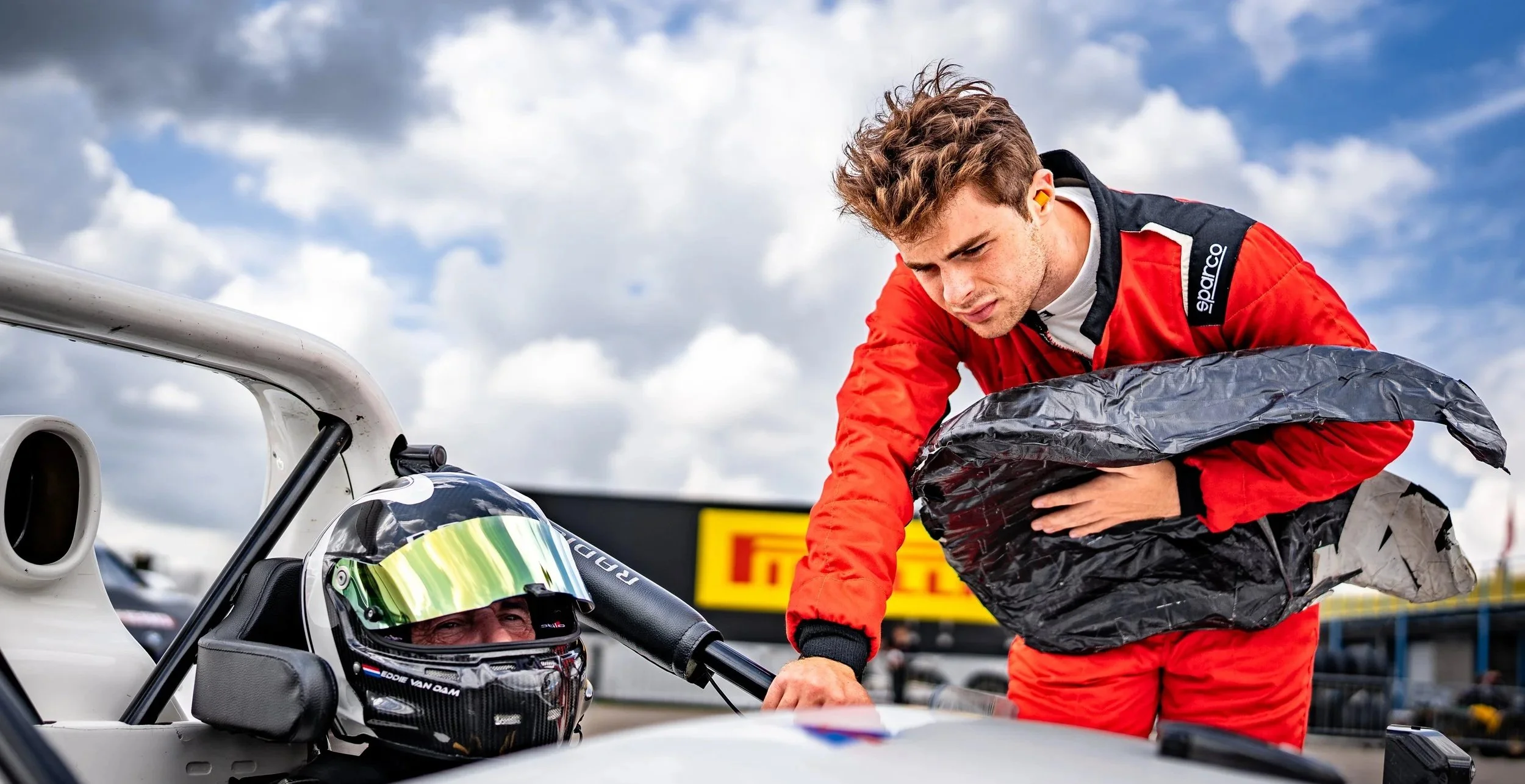A race car driver in a helmet sitting in a race car, talking to Miles Wragg in a red racing suit as they prepare for a race. The man in the suit is holding a seat and appears to be giving instructions or advice.
