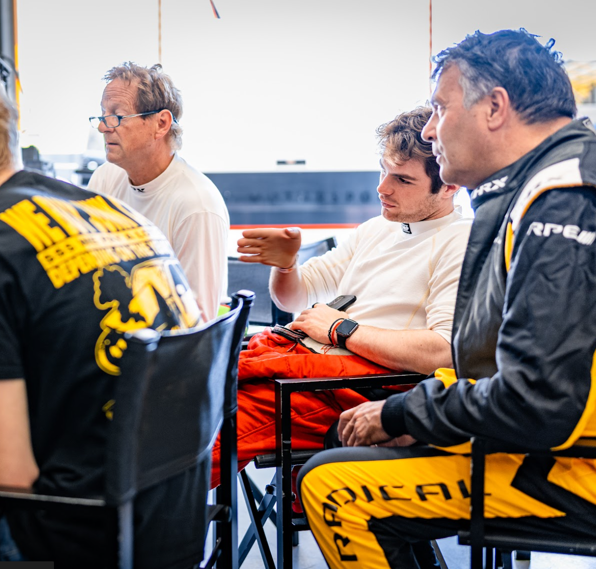 Miles Wragg talking to other drivers in racing uniforms and casual clothing sitting and talking in a team tent or garage area.