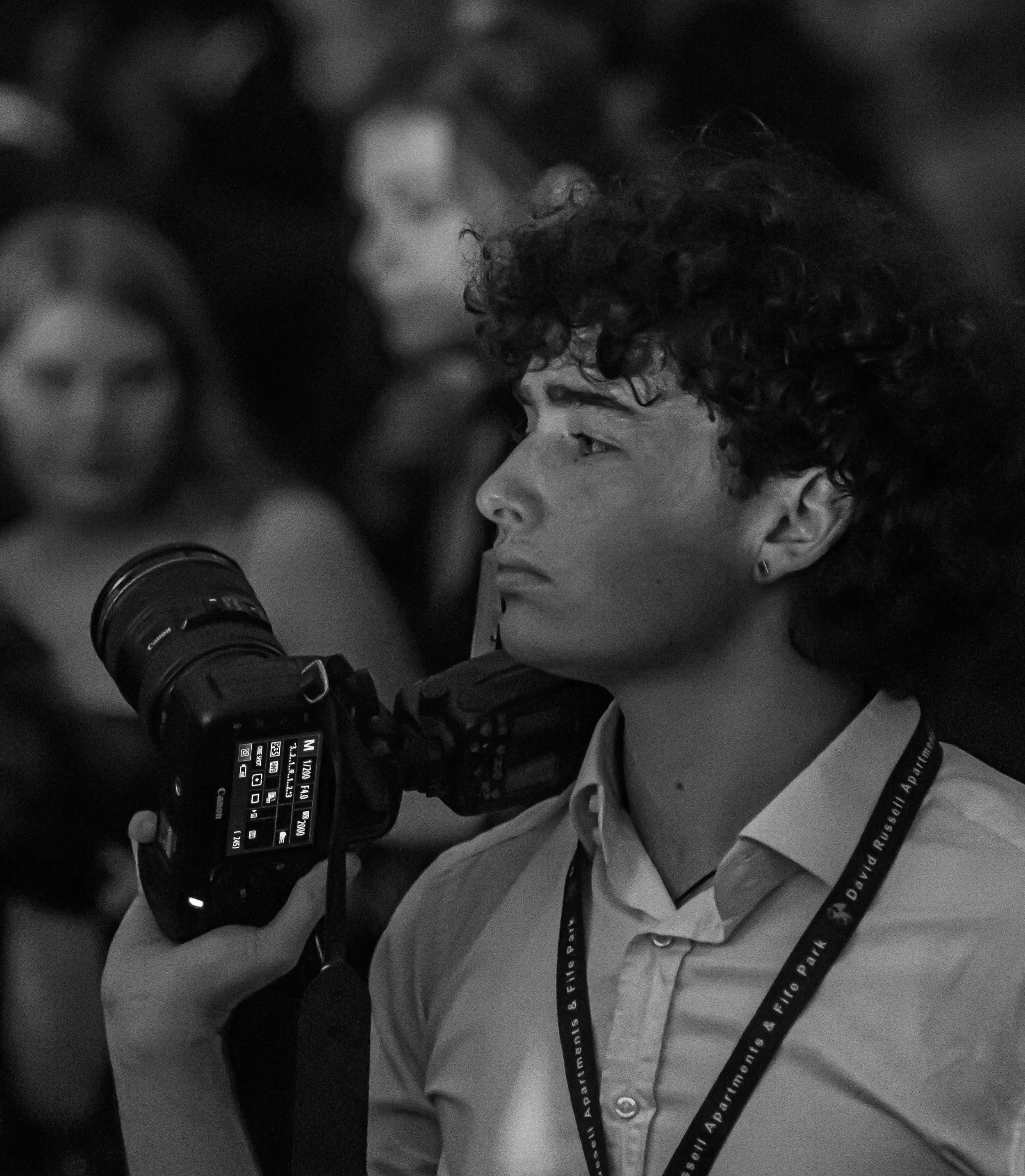 A person wearing a collared shirt with a lanyard and looking through a professional camera with a digital display screen, in black and white.