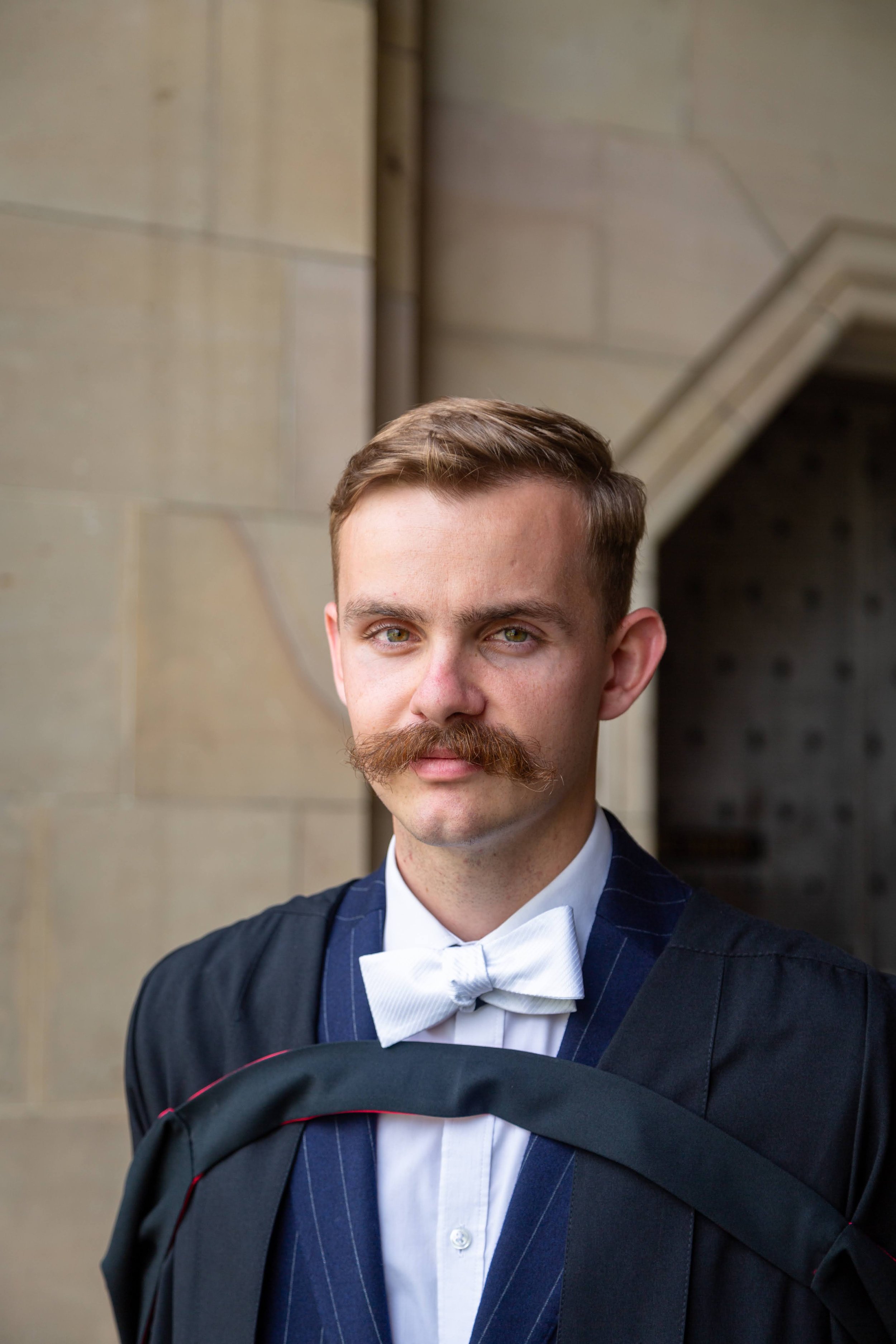 Young man with a mustache in formal attire, including a dark blue pinstripe suit, white shirt, and bow tie, standing outdoors in front of a stone building.